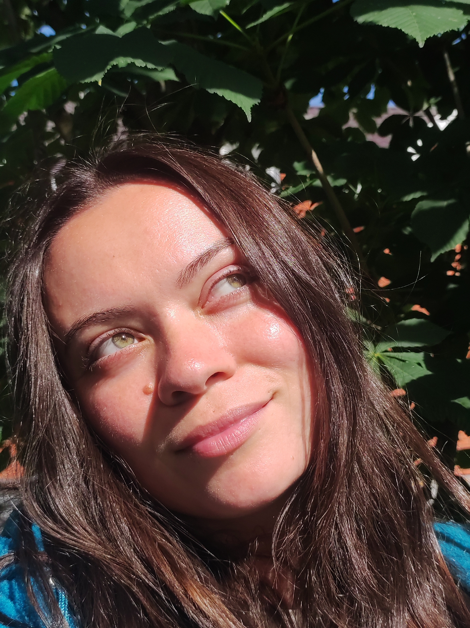 A young woman with long, dark brown hair is captured in a close-up shot, looking upwards and to the right, with a gentle smile. Sunlight dapples her face, creating highlights and shadows that emphasize her features, including her light green eyes and the soft curve of her lips. A small mole is visible on her cheekbone. She is wearing a blue garment, possibly a sweater or jacket.
The background is dominated by lush green leaves, suggesting an outdoor setting, likely in a park or garden. The dappled sunlight filtering through the foliage indicates it is daytime, possibly mid-morning or afternoon, with clear weather. The overall mood is serene and peaceful. The subject's gaze and smile convey a sense of quiet contemplation or pleasant reflection.
The image appears to be a selfie, given the close-up perspective and the way the woman is framed. The lighting and natural elements suggest a connection with nature. No text is visible in the image. The setting is implied to be in Фоки, Russia.