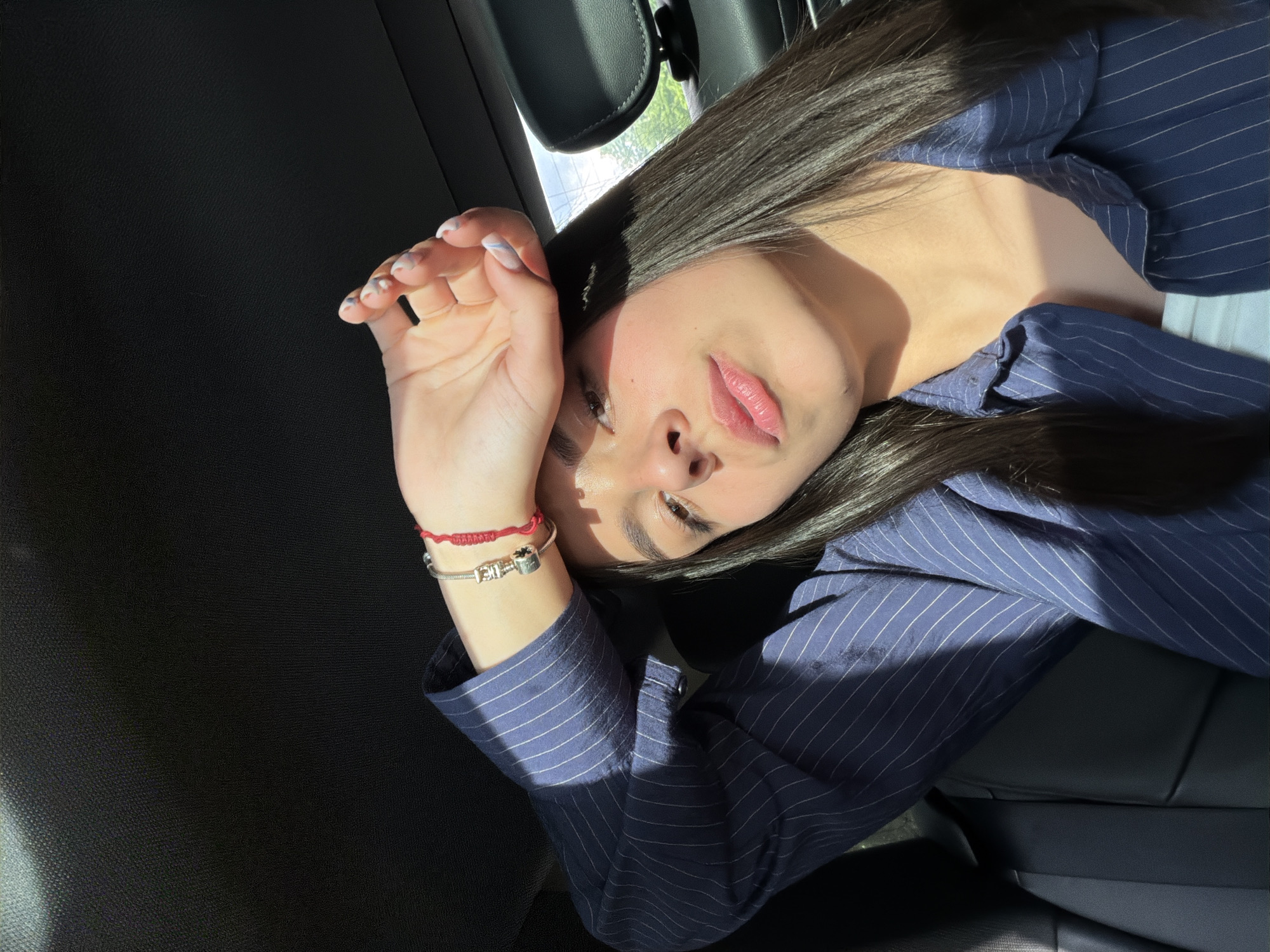 The image shows a woman lying in what appears to be the backseat of a car. The car seat and side panel are visible. The woman is wearing a striped blue shirt. She has long, dark hair. Her hand is raised near her face, possibly shielding her eyes from the sun. The location is not discernible from the image. Хаджийско, Bulgaria is not deducible from the image.