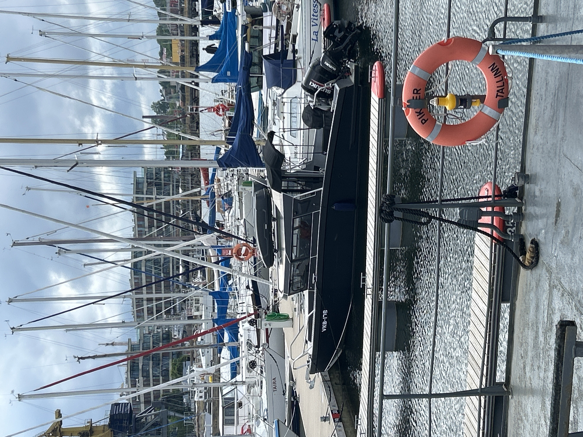 The image shows a harbor in Tallinn, Estonia, with numerous sailboats docked. In the foreground, a dark-colored motorboat with the registration VKB-178 is moored to a wooden pier. The boat has two outboard motors and a cabin. A bright orange life preserver with "PIKER TALLINN" written on it is hanging on a railing in the foreground. In the background, several buildings and cranes are visible, suggesting an urban waterfront. The sky is overcast.