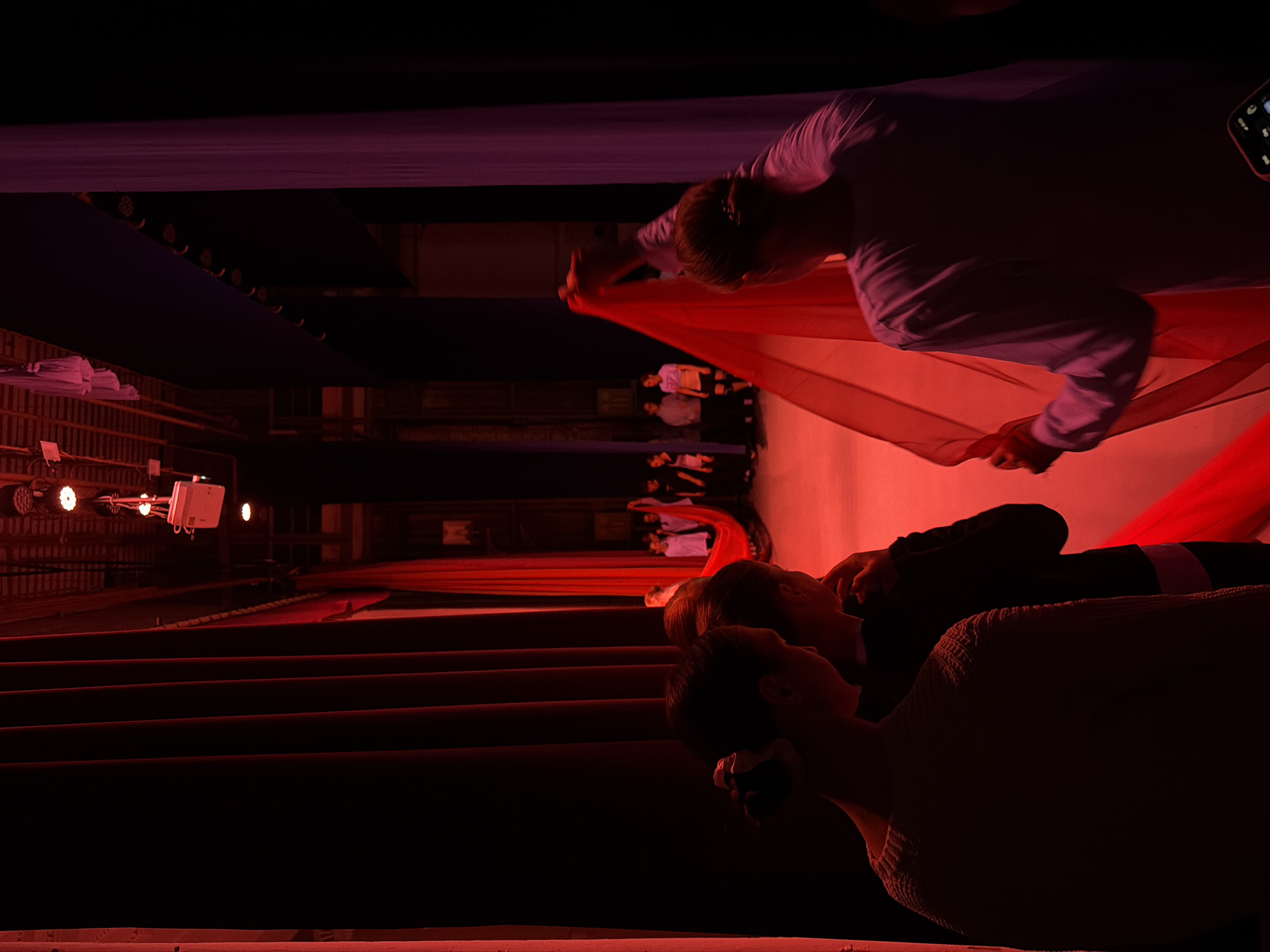 The image captures an indoor scene, likely a theater or performance hall in Perm, Russia, characterized by dramatic, low-key lighting dominated by deep reds and purples, creating an atmospheric setting.
In the foreground-midground, a person wearing a light long-sleeved shirt, seen from behind, actively manipulates a large swathe of vibrant red fabric, stretching it horizontally across the frame. Above this, another dark purple fabric is draped, contributing to the rich color palette.
To the left, receding rows of dark red auditorium seating suggest an audience area. Stage lights on stands are visible further back on the left, emitting bright, circular glows. In the background, several individuals are present on what appears to be a stage or elevated platform, some standing, some seated, dressed in casual attire, observing or participating in the event.
Closer to the bottom right, two spectators, one in a dark top and another in a light, textured sweater, are partially visible, looking towards the performance area, their features softly illuminated by the red light. A small electronic device with illuminated elements is faintly visible in the upper right. The overall atmosphere is artistic and focused, indicative of a performance, rehearsal, or fashion show. No discernible text is visible in the frame.