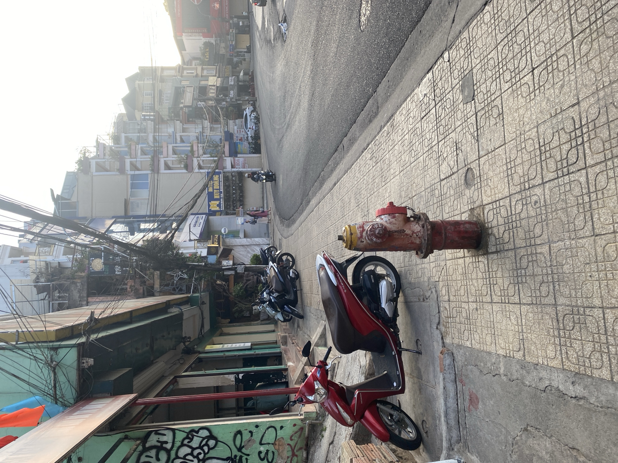 A street scene in Da Lat, Vietnam, shows a red fire hydrant on a patterned tiled sidewalk with a red Honda scooter parked next to it. Further along the sidewalk are parked motorbikes. The street, partially visible, has vehicular traffic. Buildings line the left side of the street, interspersed with overhead power lines. The scene is captured in daylight with a light-filled sky in the background.