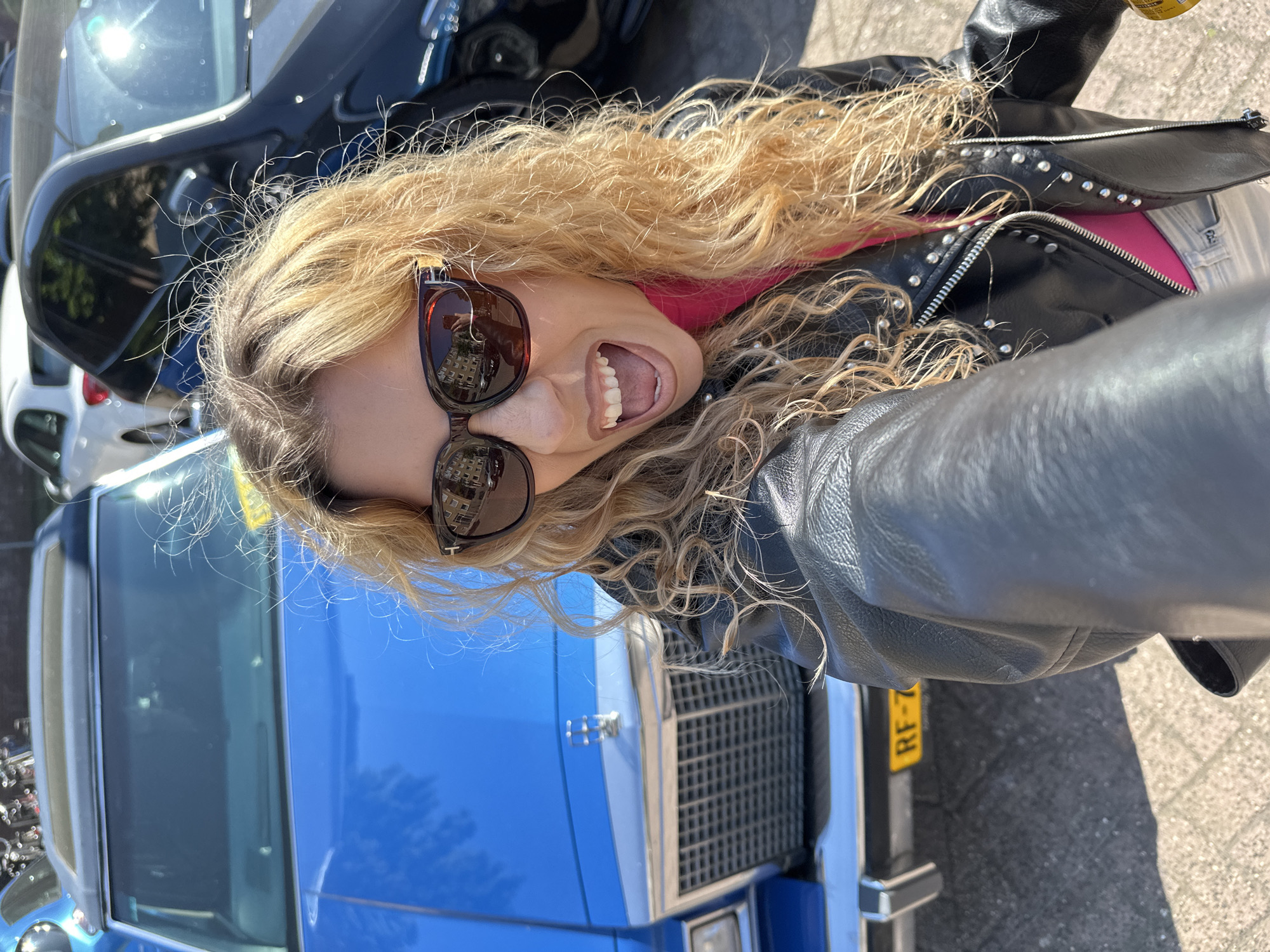 A woman with curly blonde hair, wearing sunglasses and a black leather jacket, is smiling with her mouth open in a selfie-style photo. Behind her is a blue car with a classic grille, and the license plate "RF-72" is visible. Other vehicles are visible in the background, including a black car and a white vehicle. The reflection in the woman's sunglasses shows buildings. The photo appears to be taken on a city street, and based on the license plate and architecture, it is likely taken in Amsterdam, Netherlands. The environment seems sunny.