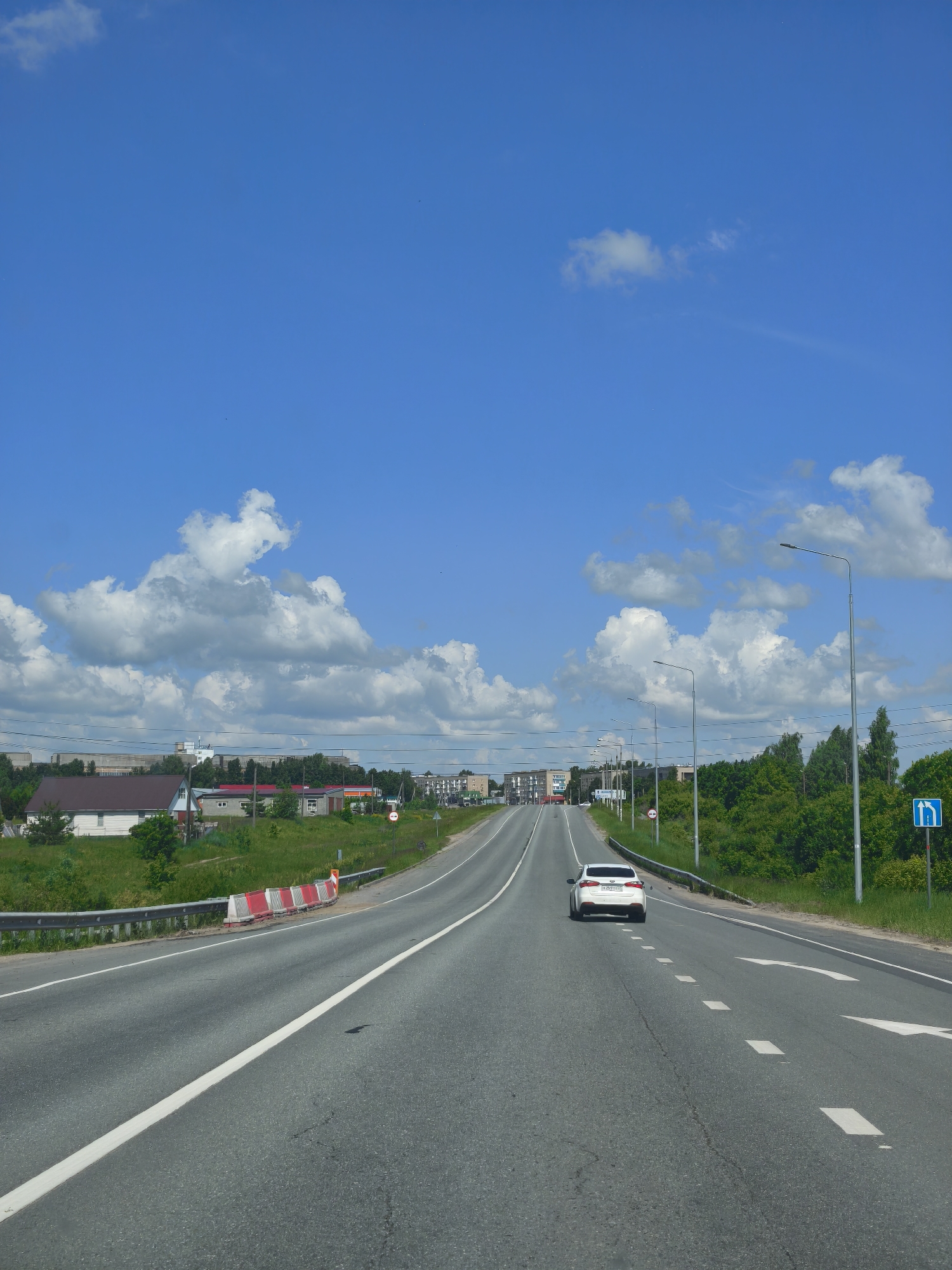 A white car drives on a multi-lane highway in Kanash, Russia, on a clear day with scattered clouds. The highway has white lane markings and a guardrail on the side. Adjacent to the highway is a grassy verge with trees and some buildings in the distance, including houses and what appear to be industrial buildings. A blue road sign indicating a turn is visible. There are streetlights along the road and traffic cones further up the highway.