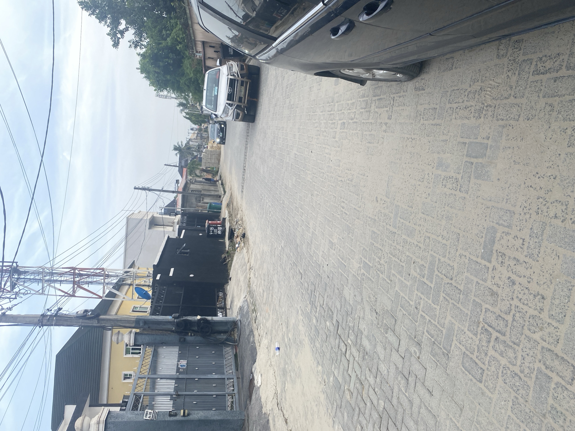 The image shows a street in an urban environment, likely residential. The road is paved with light grey paving stones laid in a herringbone pattern. A dark colored car is parked on the right side of the frame, with only the side and a portion of a tire visible. Further down the street, two light-colored vehicles are parked, one appearing to be an SUV. On the left side of the street, there are buildings, some with gated entrances. Power lines and a communication tower are visible in the background. The sky is overcast and the overall scene is bathed in muted daylight.