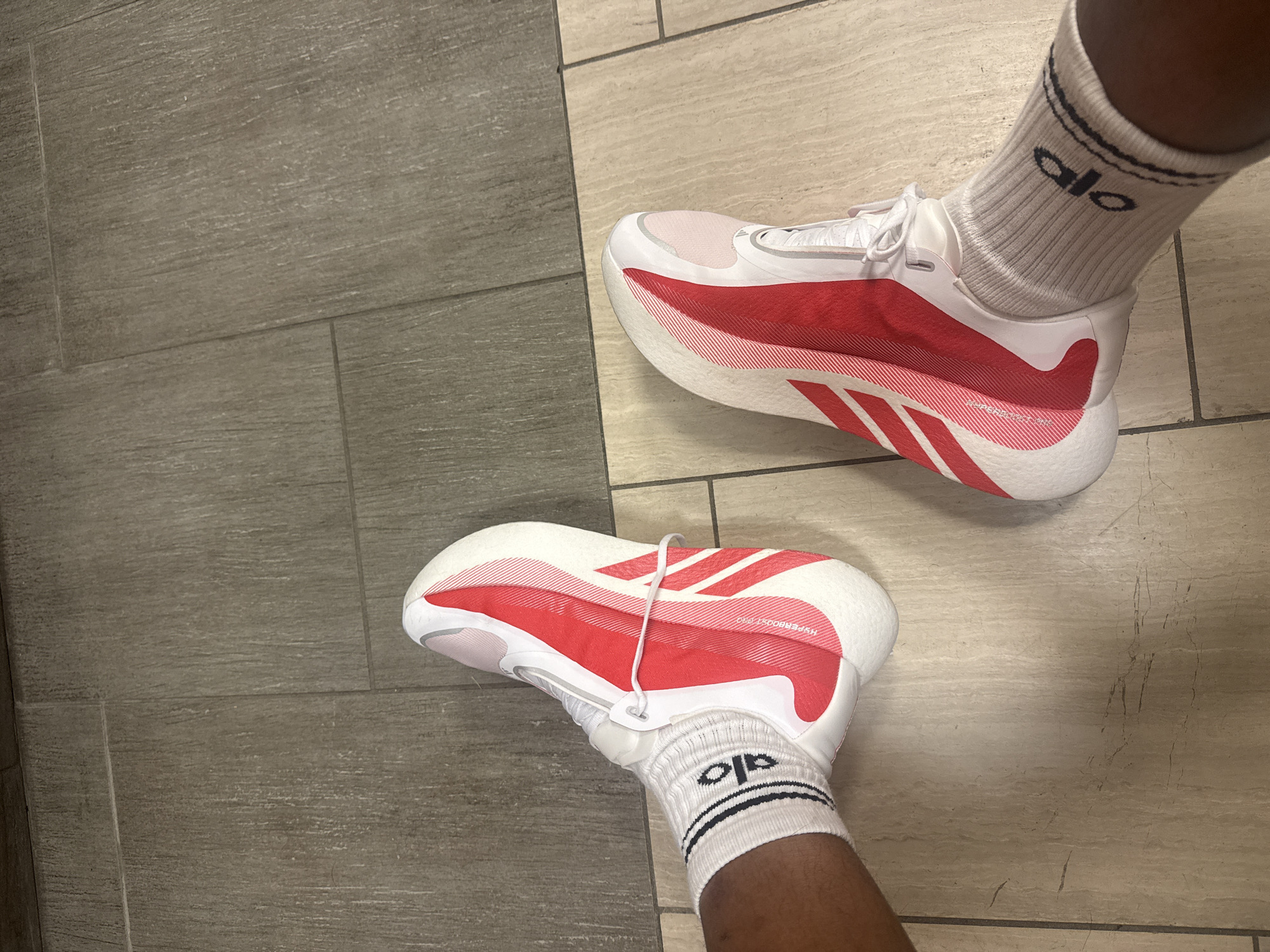 The image shows a top-down view of two feet wearing white socks with the "alo" logo, and white running shoes with red accents. The shoes appear to be Adidas Hyperboost Pro model. The feet are standing on a tiled floor with a gray and tan pattern. The photo was likely taken indoors. The location is unknown, but is in Los Angeles, USA.
