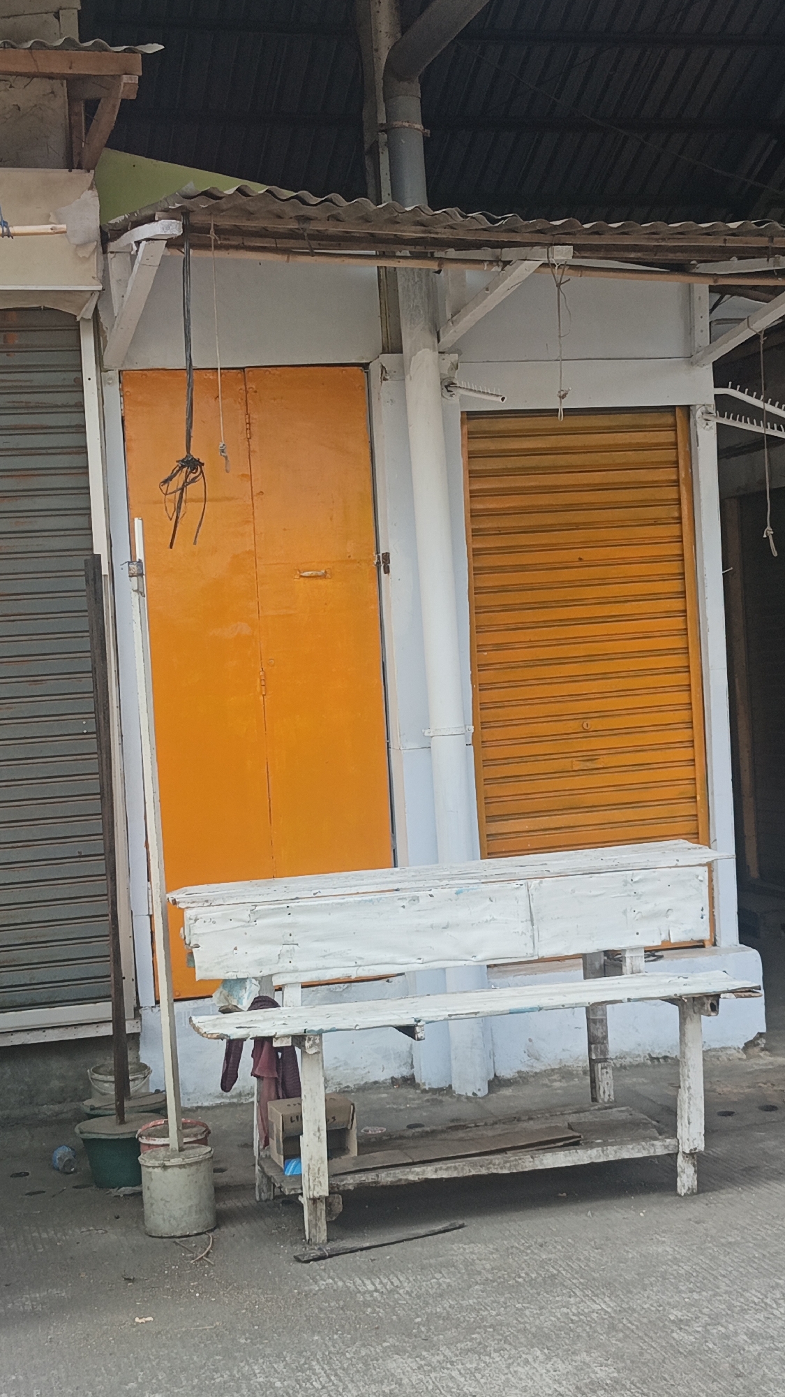 The image depicts the exterior of a small, shuttered shop in Jekulo, Indonesia. The shop has two prominent orange roll-up doors, one larger than the other. In front of the shop is a weathered, white wooden bench with a lower shelf, currently empty except for a small cardboard box and some debris. To the left of the bench, there are several buckets, one green and two gray, with a pipe or pole standing upright next to them. The shop itself appears to be constructed with a light-colored outer wall and a corrugated metal roof supported by wooden beams. The overall scene suggests a quiet, perhaps abandoned or closed business in a local setting.