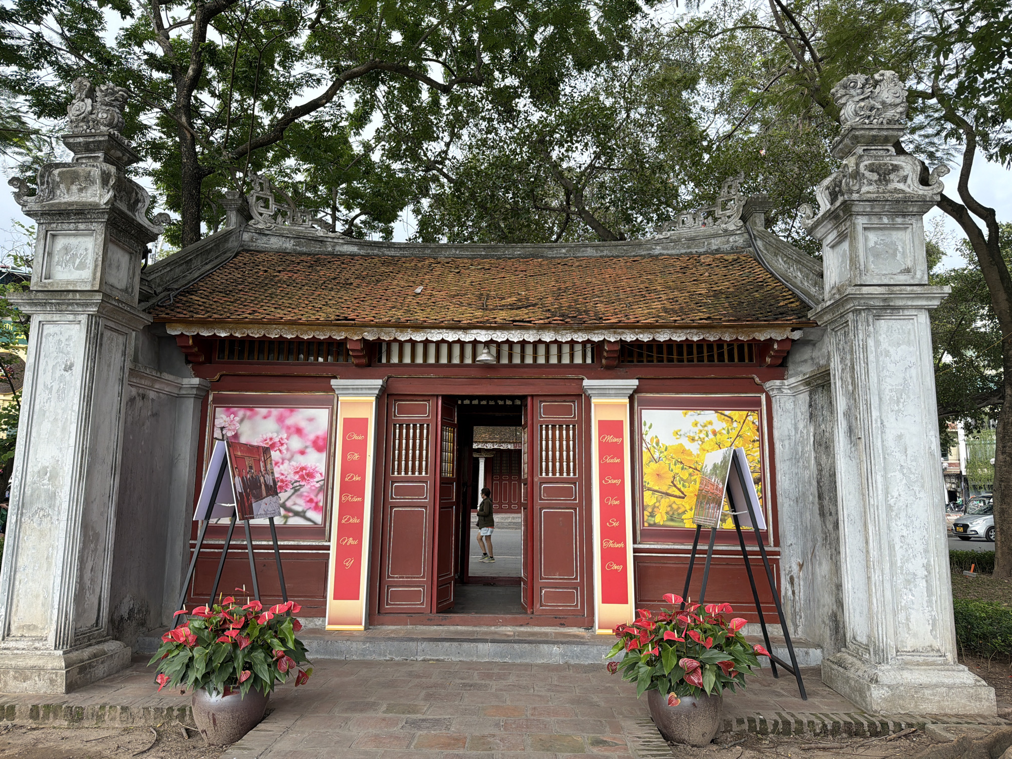 The image depicts an entrance to a traditional Vietnamese structure in Hà Nội, Vietnam. The building has a red-tiled roof with decorative elements along the roofline. The main entrance consists of a set of red wooden doors, which are open, revealing an inner courtyard or room.
Flanking the entrance are two large stone pillars, each topped with a sculptural element. Next to each pillar, there is a display of flowers in a pot, along with a framed poster or artwork supported by an easel. The poster on the left features pink blossoms, while the poster on the right displays yellow flowers. Banners with vertical text, in Vietnamese script, are placed between the doorways and the displays.
A person stands in the doorway, partially obscured by the red doors and the interior of the structure. The area in front of the building is paved with brick or stone. Above the building, trees with green foliage are visible, creating a canopy over the scene.