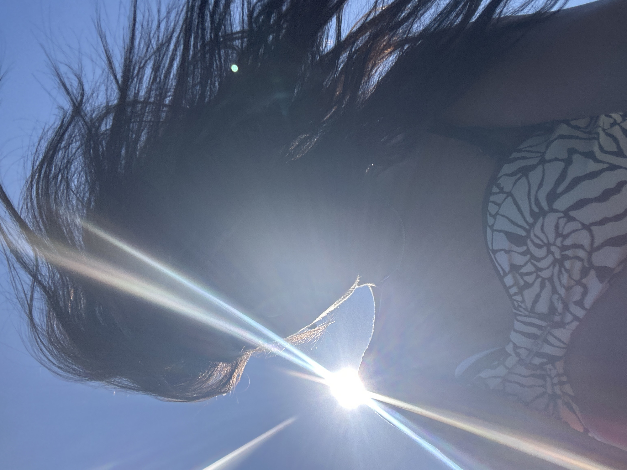 A low-angle, close-up shot captures a person from the shoulders up, silhouetted against a bright blue sky and the sun. The sun is positioned prominently to the left of the frame, creating a strong lens flare with visible light rays emanating outwards. The person's long, dark hair is blown by the wind, obscuring much of their face, though a portion of their profile is visible. They are wearing a patterned bikini top with black and white markings that resemble zebra stripes or abstract floral shapes. The scene is set outdoors with a clear sky, suggestive of a sunny day. The provided location of Cala Lliteres, Spain, indicates a coastal or beach environment.