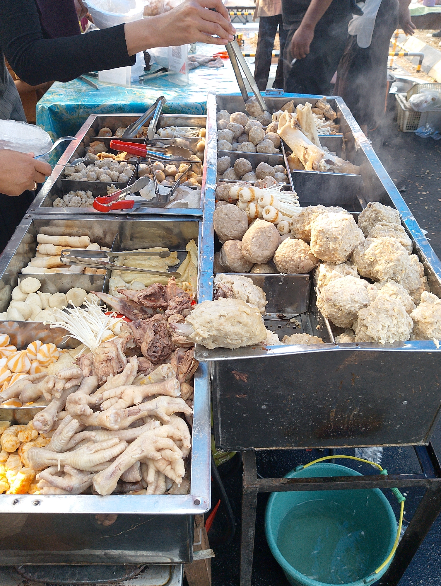 This is a close-up, eye-level shot of a food stall, likely located in Karawang, Indonesia, showcasing an array of prepared food items. The scene is dominated by several large, stainless steel containers filled with various ingredients, suggesting a preparation or serving area for a meal.
In the foreground, the leftmost container is filled with raw chicken feet, which are pale and appear to have been prepped. To the right of the chicken feet, a container holds white enoki mushrooms. Above the mushrooms, there are pieces of cooked chicken meat, some appearing to be bone-in. Further to the right, a large container is filled with light-colored, spherical meatballs or fish balls, with some steam rising from them, indicating they are hot. Another container to the left holds a mixture of white, circular food items and what appear to be pieces of processed tofu or fish cake, some of which are striped orange and white. Tongs are visible in several containers, suggesting active preparation or serving.
In the background, the scene transitions to a blue and white patterned tablecloth. Several pairs of hands are visible, actively engaged with the food. One person, on the left, is holding a pair of tongs over a container of food. Another person, in the upper center, is holding tongs and reaching towards a container. The clothing of these individuals is partially visible, including dark sleeves and lighter fabric. The overall impression is of a bustling food stall with a variety of ingredients ready to be served. A turquoise bucket filled with water is visible at the bottom right of the frame, underneath the food containers.