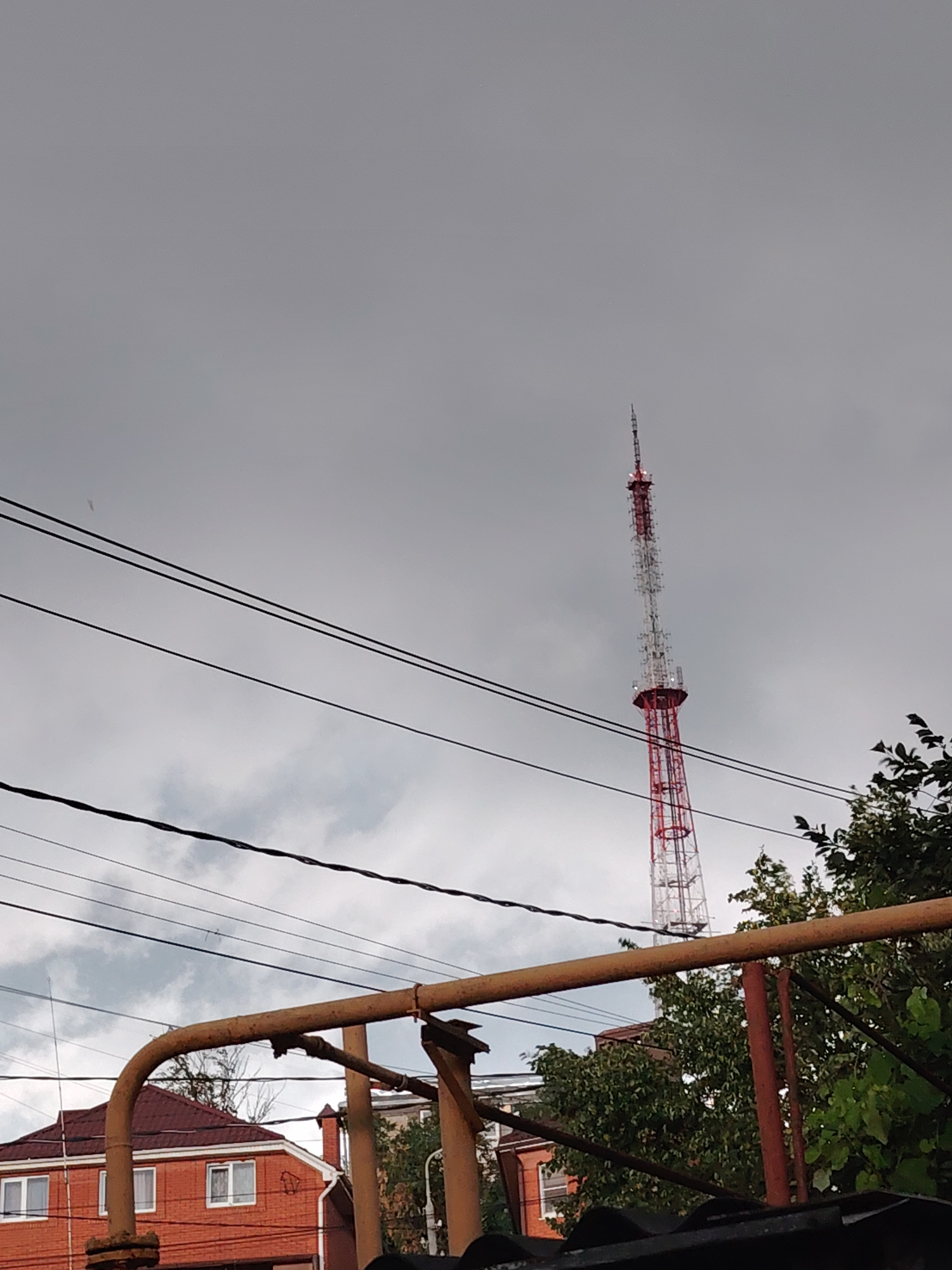The image shows a red and white television tower in the distance, with a cloudy sky. In the foreground, there is a rusty metal structure, power lines, and some greenery. The lower left of the image shows a red brick house with white-framed windows and a dark red roof. The scene appears to be in an urban or suburban setting.