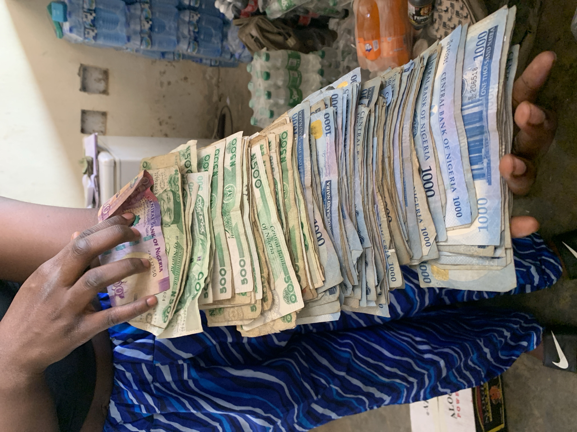 A person sits wearing blue patterned trousers, holding a large wad of Nigerian Naira banknotes. The denominations include ₦500 and ₦1000 notes. The background shows stacked bottled water and beverage bottles. The location is suggested to be in Kabong, Nigeria, based on the currency and implied context. The scene depicts a person holding a substantial amount of cash.