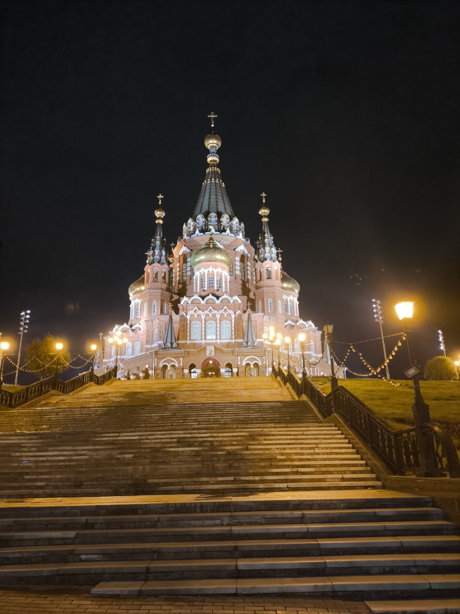 The image displays the illuminated St. Michael's Cathedral (Mikhailovsky Cathedral) in Izhevsk, Russia, at night. The cathedral, constructed primarily of reddish-brown brick, features multiple domes, spires, and intricate architectural details. The central dome is large and dark-colored, topped with a golden orb and a cross, flanked by smaller golden domes and spires, each surmounted by a cross. Numerous windows with white frames are visible across the facade.
A wide, multi-tiered stone staircase dominates the foreground, leading directly up to the main entrance of the cathedral. The stairs and the lower parts of the cathedral are brightly lit by numerous warm-toned streetlights mounted on decorative black poles positioned along the sides of the staircase and within the church grounds. A dark metal railing runs alongside the stairs.
On either side of the staircase, grassy slopes are visible, also illuminated by the streetlights, giving the grass a warm, golden hue. String lights with circular bulbs are strung between poles along the right side of the stairs. In the mid-ground, on both the left and right sides of the frame, sets of tall poles with multiple floodlights are visible, further illuminating the area. The sky above is dark and cloudless, characteristic of a clear night. No individuals are present in the scene.