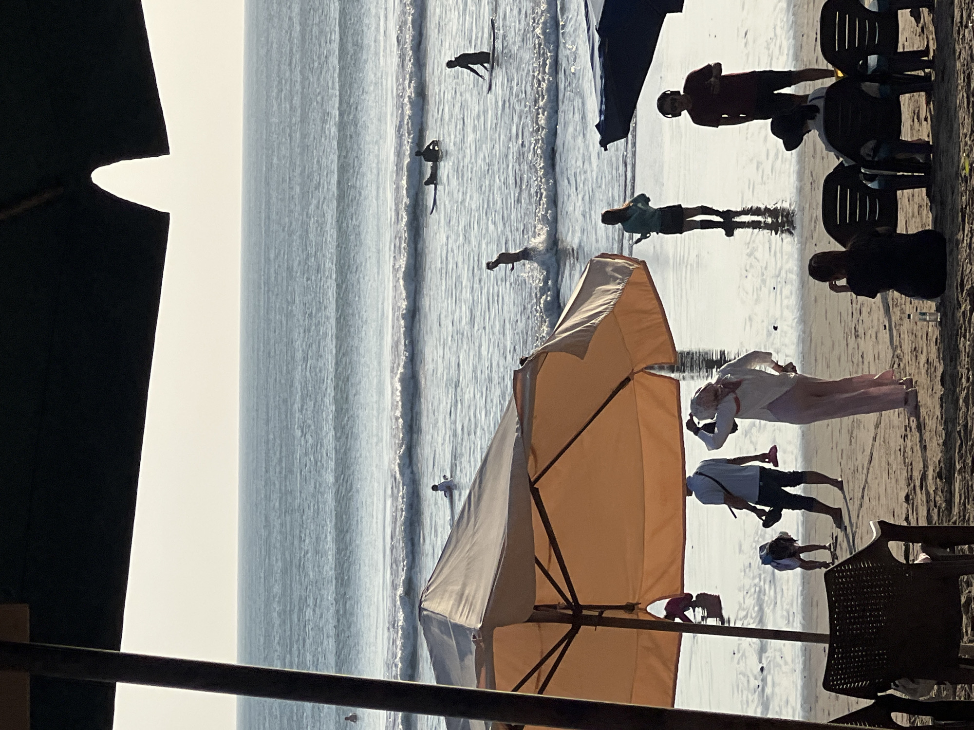 The image depicts a serene beach scene at Kuta, Indonesia, bathed in the warm glow of the late afternoon sun. The ocean, a tranquil expanse of blue, meets the shore with gentle waves. Several surfers are scattered across the water, some paddling on their boards, others waiting for the perfect wave.
On the wet sand near the water's edge, a diverse group of people are engaged in various activities. A woman in a hijab and a long, flowy outfit stands near the center, her back to the camera, appearing to be taking a picture or adjusting her attire. To her left, a man in a blue shirt and dark pants holds a pair of flip-flops. A child in a white shirt and a hat is bent over, seemingly playing in the sand.
Further back, silhouetted against the bright sky, a woman in a teal top and black skirt stands, holding something in her hand. Near her, a man in a red t-shirt and dark shorts, wearing headphones, looks down at his phone. In the foreground, three chairs are occupied by individuals, their backs to the viewer, suggesting they are either resting or observing the scene.
The beach is dotted with umbrellas, providing shade and adding to the relaxed atmosphere. The overall impression is one of a peaceful day at the beach, with a mix of leisure activities and people enjoying the coastal environment of Kuta.