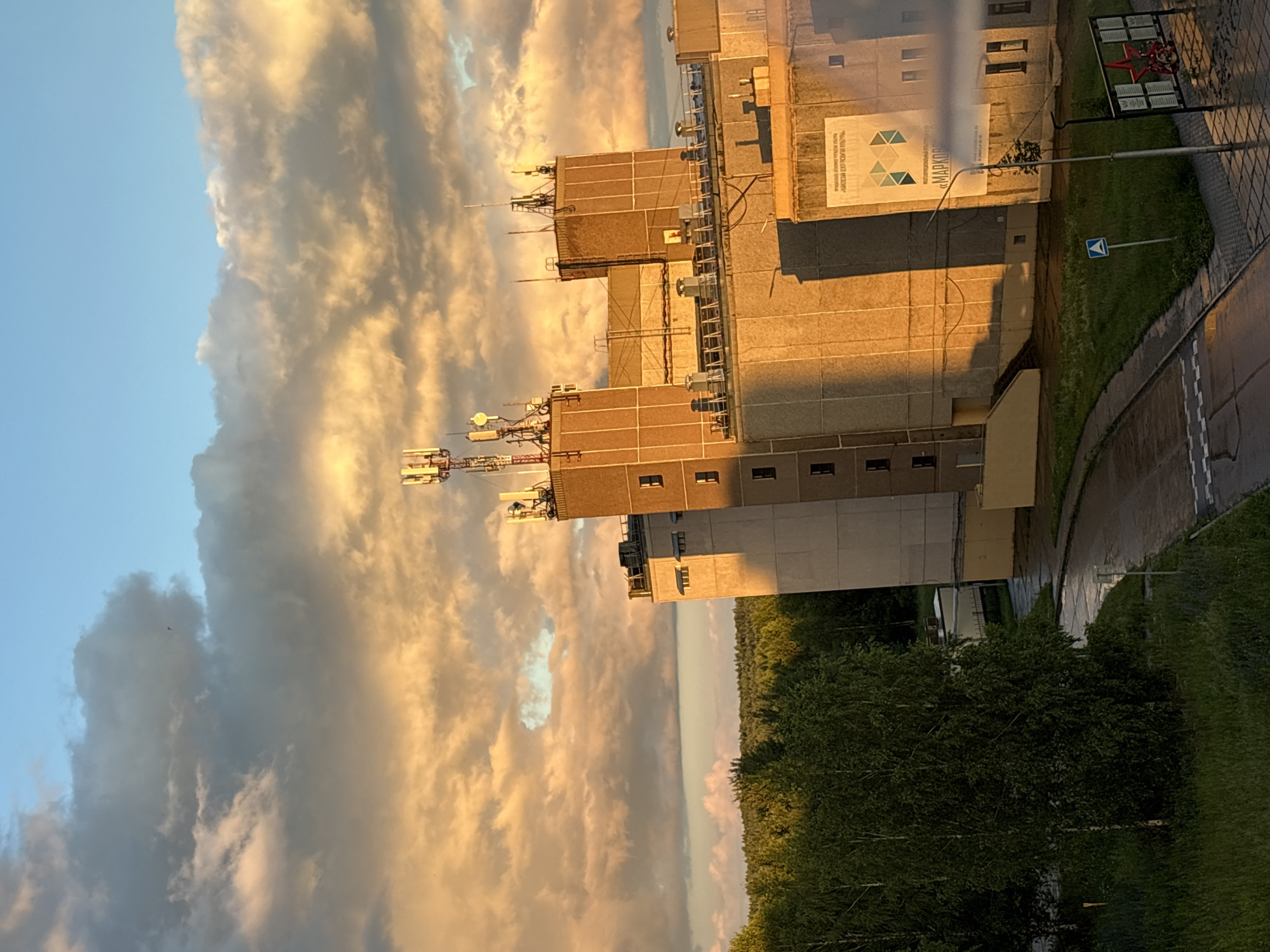 The image depicts a scene in Марковский, Russia, likely during sunset due to the warm, golden light illuminating the clouds and buildings. The sky is filled with dramatic, voluminous clouds, some tinged with orange and yellow from the setting sun, while others appear darker and more gray. The primary objects in the foreground are several multi-story buildings, appearing to be industrial or office structures. These buildings have visible antennas and communication equipment mounted on their roofs, suggesting a possible telecommunications hub or research facility.
The scene includes a paved area that appears to be a road or pathway next to a grassy strip. There are trees visible in the background, adding a natural element to the otherwise man-made landscape. The lighting suggests it is late afternoon or early evening.
A notable detail is a sign on one of the buildings that reads "МАРКОВ" in large, stylized letters, and below it, smaller text, likely a company name or slogan, though it is difficult to read clearly. There is also a red star emblem mounted on a post, which could be a historical or memorial marker, common in Russia. The overall mood is somewhat stark but also serene, with the dramatic sky providing a visually striking backdrop to the functional architecture. No people are visible in the frame.