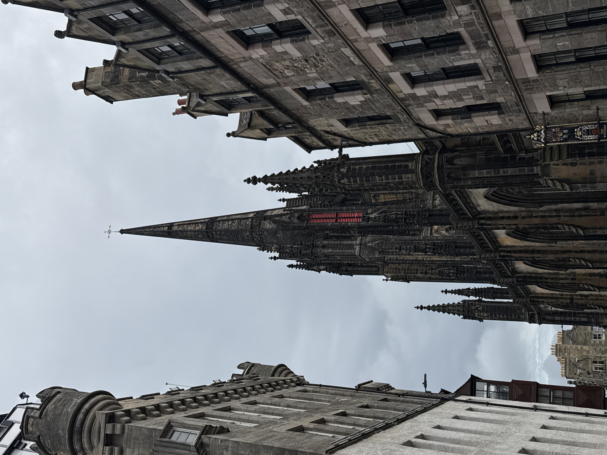 The image captures a view looking up at the Victoria Street in the city of Edinburgh, United Kingdom. On the left, a building with a rounded turret and crenellated battlements is visible. To the right of the turret, the ornate Gothic spire of the Tolbooth Kirk dominates the skyline. This spire features intricate stonework, flying buttresses, and a cross at its apex. A section of the spire exhibits bright red stained glass. Flanking the spire on the right is a building constructed of grey stone, characterized by multiple windows with dark frames, and punctuated by chimneys. The sky above is overcast and grey.