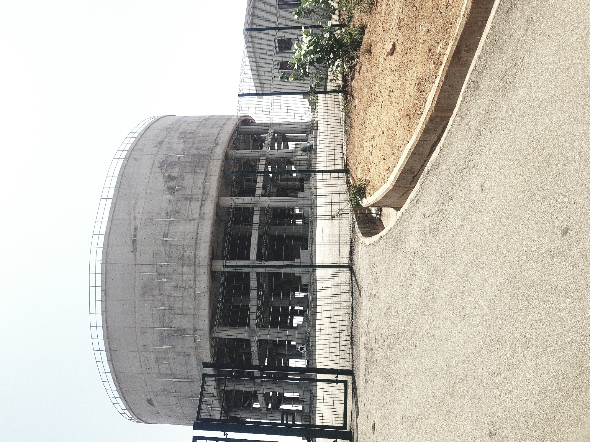 The image shows a concrete cylindrical structure under construction, possibly a water tower, located in Bauchi, Nigeria. The structure has a metal framework and support pillars visible through its open design. A wire fence surrounds the base of the structure, and a paved road runs alongside it. A building with windows is visible in the background, and an area of dirt and vegetation is adjacent to the road. The sky is overcast.