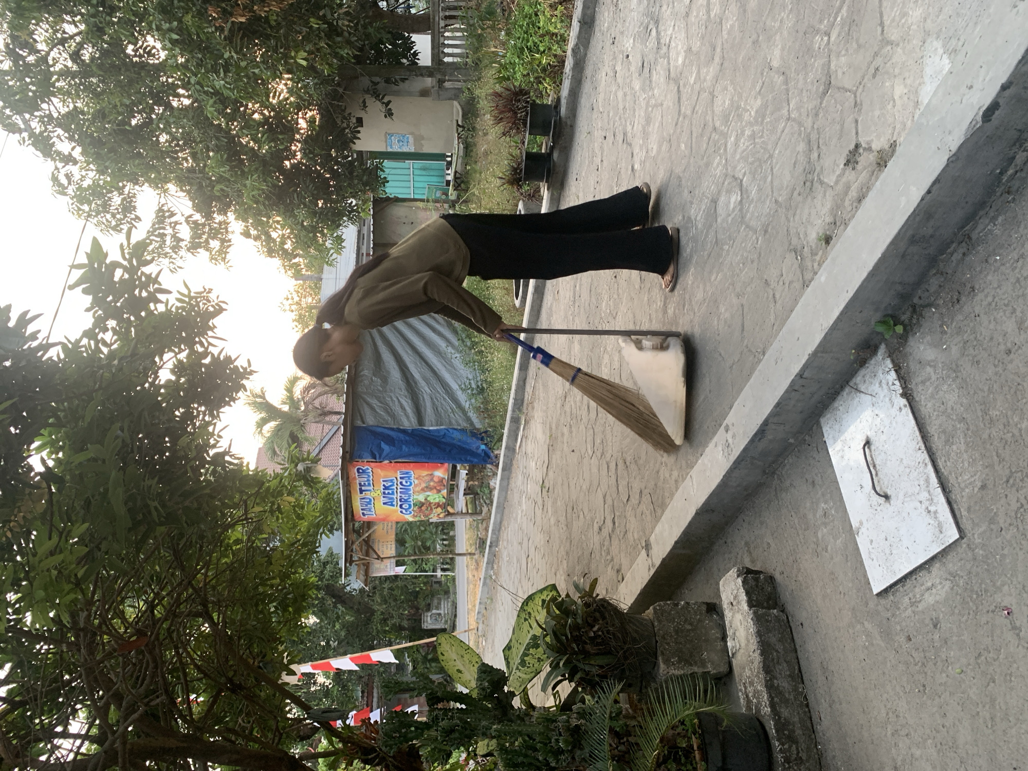 In the city of Sumberjo, Indonesia, a young person with dark hair tied back is sweeping a concrete path with a broom and dustpan. They are wearing a long-sleeved olive green shirt and black pants, with their feet in flip-flops. The path appears to be outdoors, possibly in a residential or commercial area, given the presence of trees, plants in pots, and a building in the background. A banner with Indonesian text and images of food is visible on the building, suggesting a food stall or restaurant. The overall lighting suggests it is either early morning or late afternoon. The environment is somewhat rustic, with a mix of natural elements and man-made structures.