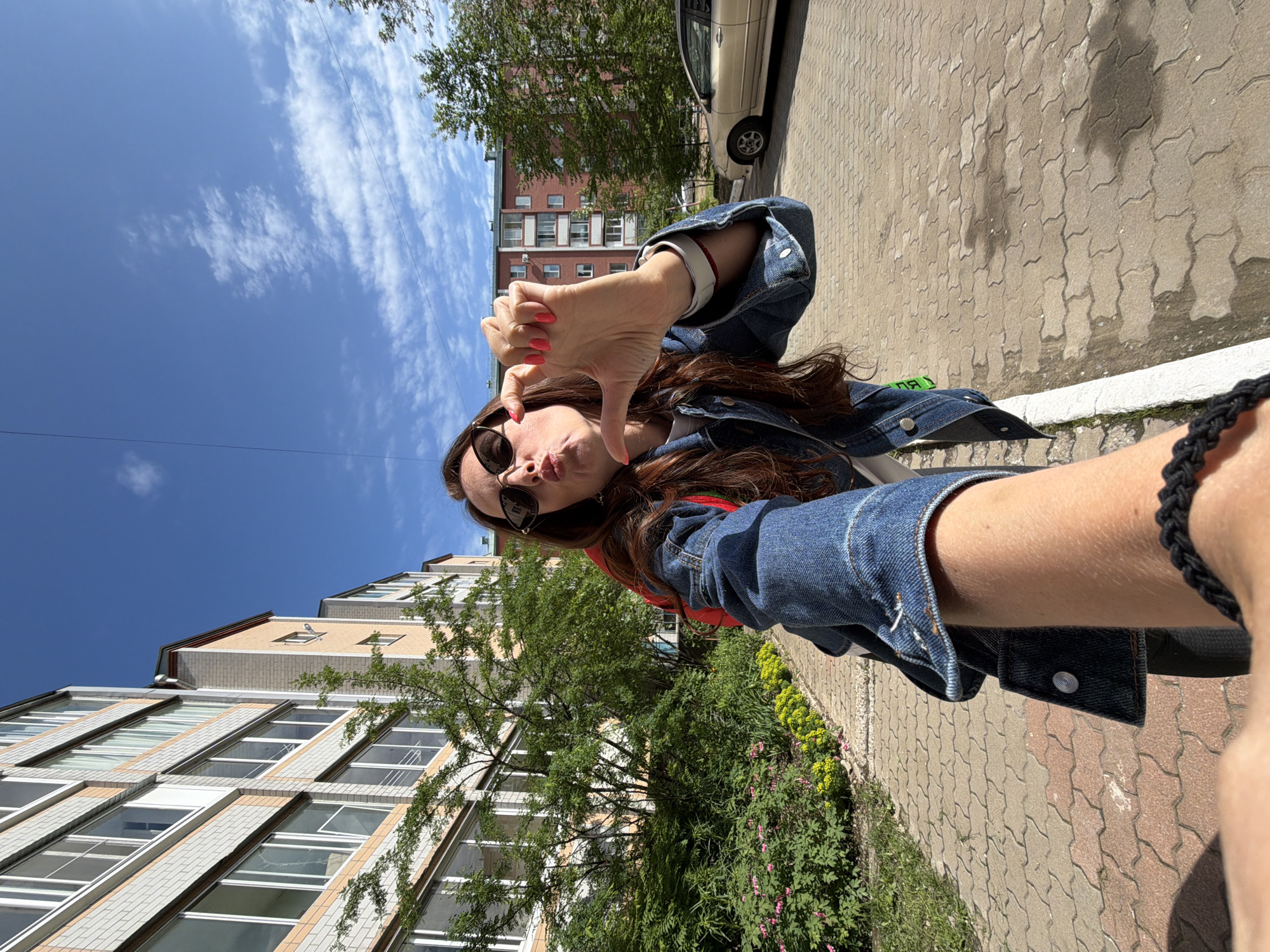 A woman with long brown hair, sunglasses, and a denim jacket poses for a selfie outdoors. She is making a "heart" shape with her fingers and puckering her lips. The background shows a multi-story apartment building, trees, and a paved walkway. A tan sedan is parked on the right side of the image. The scene appears to be in Birobidzhan, Russia, given the architectural style of the buildings and the overall environment. The sky is blue with scattered white clouds.