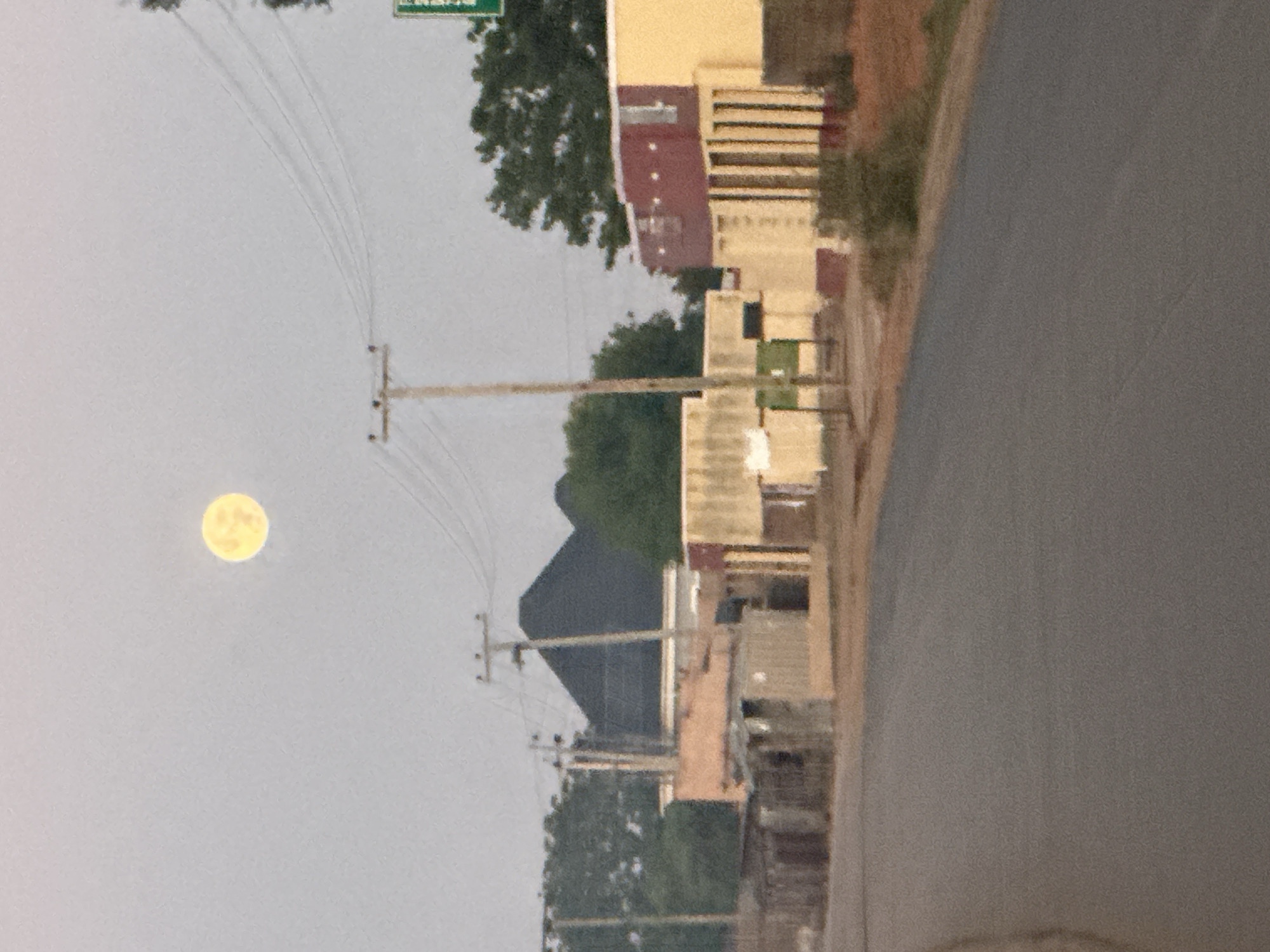 The image shows a street scene in Bukuru, Nigeria. A paved road runs along the right edge of the frame. Buildings of varying heights and colors line the street, including structures in shades of tan, red, and brown. Utility poles with wires are visible extending from the bottom left towards the upper left of the image. In the sky, a bright, yellow moon is visible. The sky is overcast, appearing light gray. Green foliage from trees is interspersed among the buildings. A green street sign is visible.