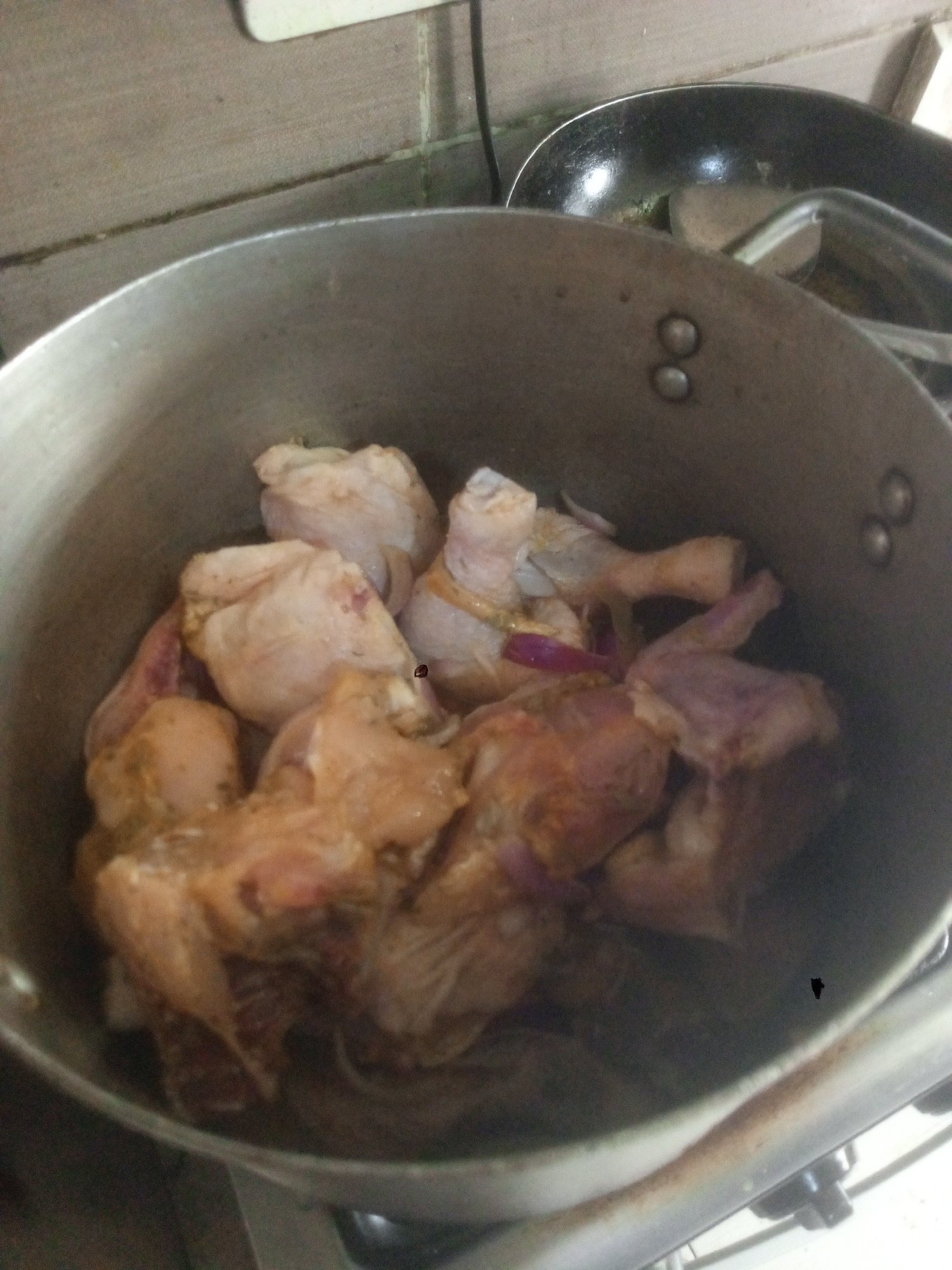 An eye-level, medium shot depicts a pot filled with raw chicken pieces on a stovetop, situated indoors. The pot is likely made of aluminum or stainless steel due to its silver-grey color. Some of the chicken pieces appear seasoned or marinated. A dark frying pan with a spatula resting inside is partially visible in the upper right corner. The background includes a tiled wall. Based on the context provided, the location is Gwarinpa, Nigeria. There is no evidence of people in the image.