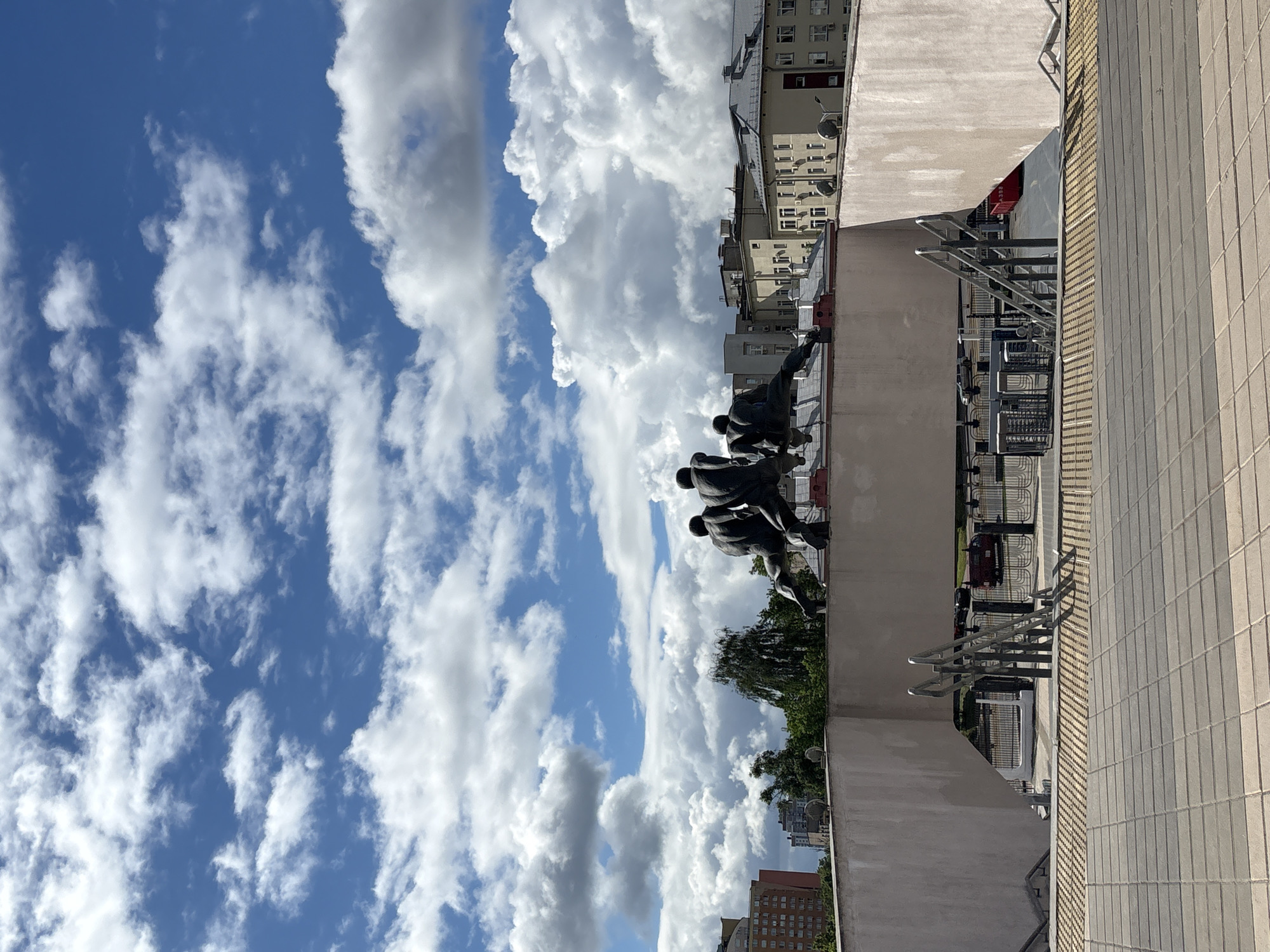 The image shows a partial view of the Minsk Hero City Obelisk in Minsk, Belarus, set against a sky filled with cumulus clouds. The scene captures a portion of the monument featuring bronze statues of figures, positioned on a concrete structure with a tiled exterior. In the background, buildings are visible, with urban greenery filling some spaces. Metal railings and what appear to be shopping carts are also visible on the edge of the rooftop.
