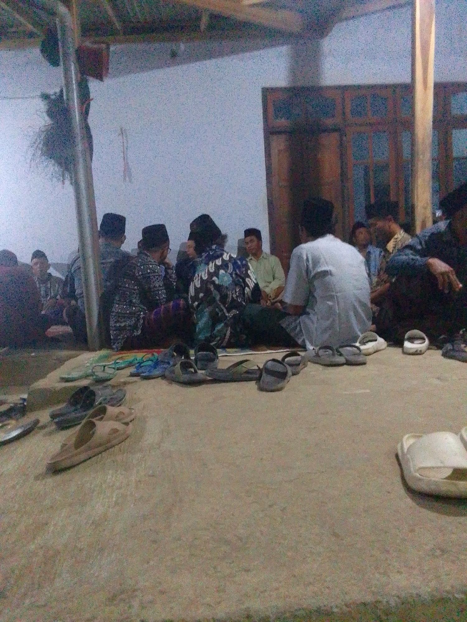In Ngares, Indonesia, a group of men are gathered indoors, seated on the floor. The room appears to be a communal space, possibly a prayer room or a meeting hall, with a thatched roof visible overhead and wooden doors in the background. Most of the men are wearing traditional Islamic attire, including peci caps, and some have their backs to the camera. A variety of footwear, including sandals and flip-flops, are scattered on the floor around the seated individuals, suggesting a casual or religious gathering where shoes are removed before entering. The lighting is dim, creating a somewhat hazy atmosphere. The overall impression is one of a community coming together for a shared activity.