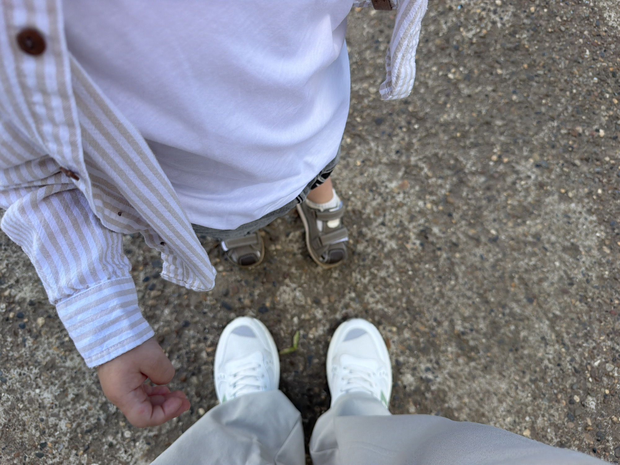 The image shows a close-up, overhead view of the lower body and arms of two individuals, a child and an adult, standing on a concrete surface. The child, on the right, wears a white t-shirt and a striped button-up shirt with beige and white stripes and two brown buttons visible. The child's left arm and hand are visible, resting on their lap. The child is wearing two small, greyish-brown sandals with white straps. The adult, on the left, wears light grey trousers and white sneakers with grey accents and white laces. The concrete surface appears to be an outdoor path or sidewalk, with visible pebbles and texture. There are no specific landmarks or indications within the image to pinpoint a location in Chaikovsky, Russia.