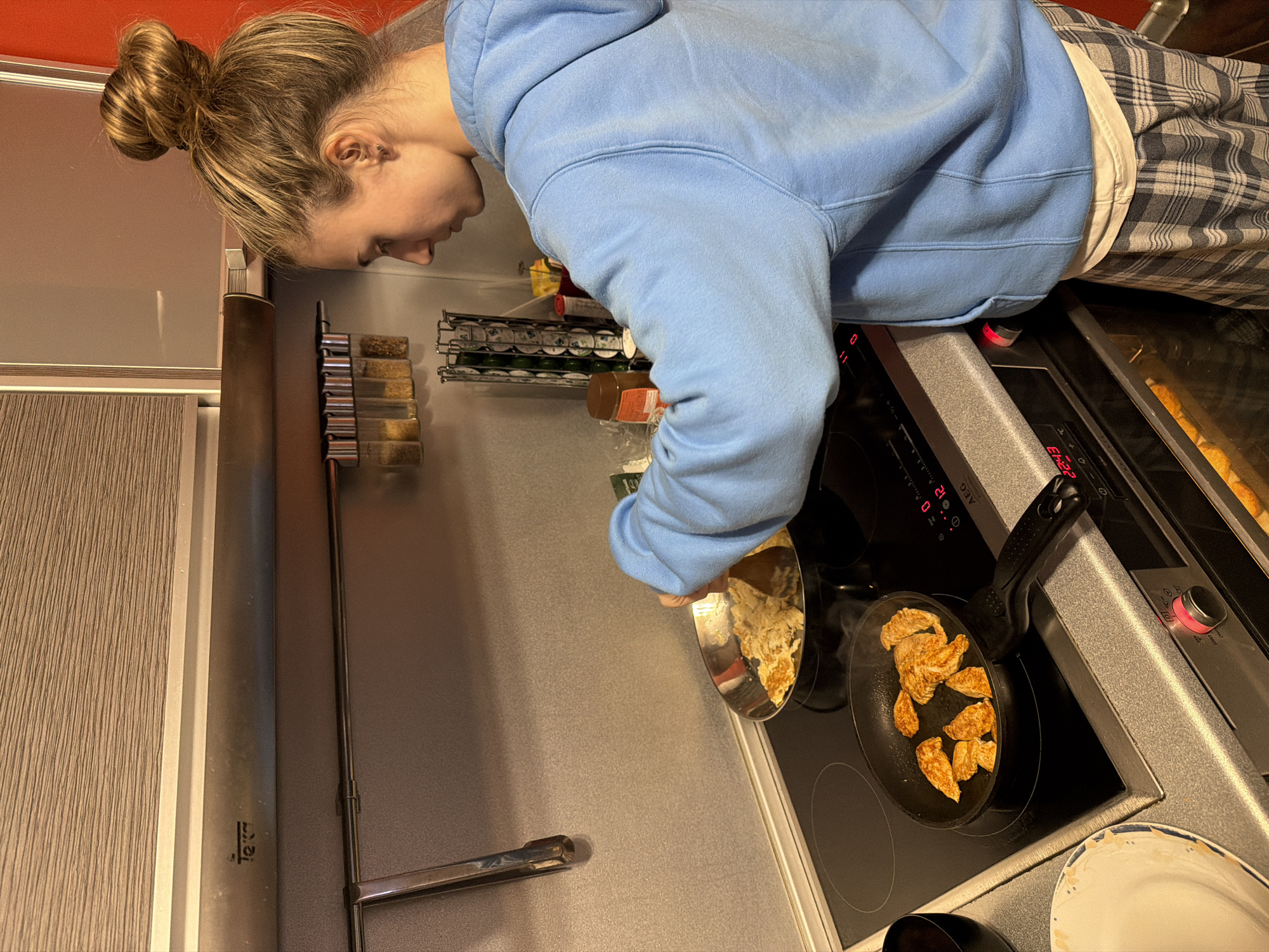 A woman with her hair in a bun is cooking in a kitchen. She is wearing a light blue hoodie and plaid pajama pants. She is using a frying pan to cook what appears to be chicken pieces. There is another pan nearby with some other cooked items. The kitchen has a modern aesthetic, with gray cabinets and countertops, and stainless steel appliances. There are spices on a rack visible in the background. The location is not possible to determine from the image.