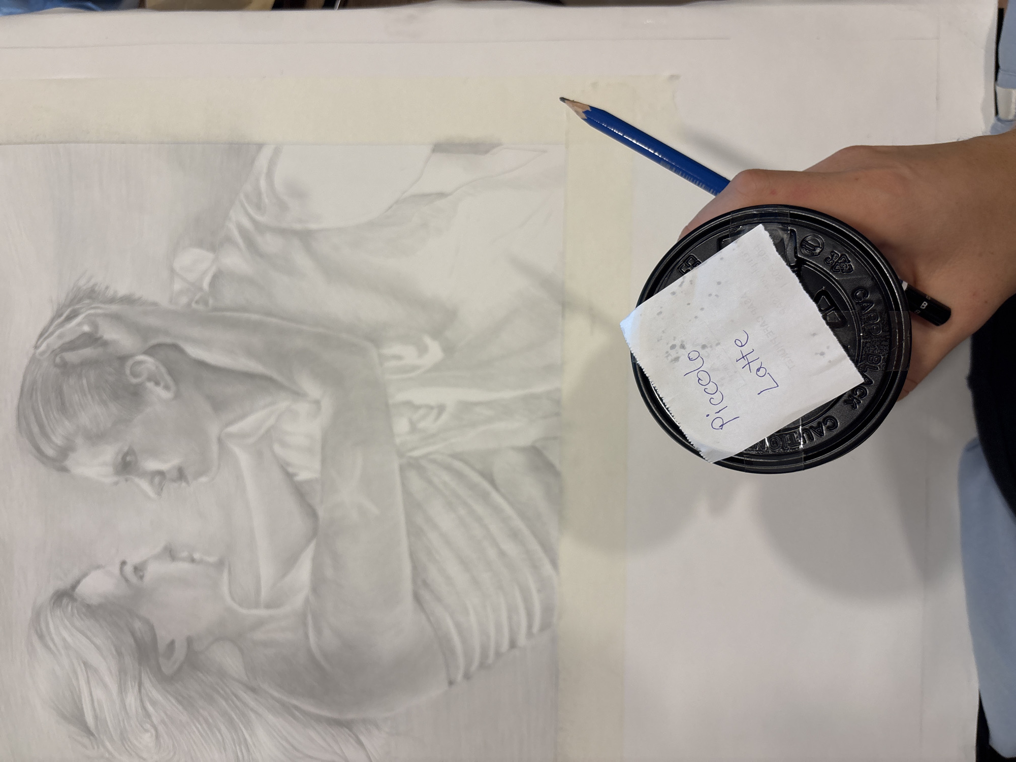 An artist in Choeng Thale, Thailand, is captured mid-activity, working on a detailed graphite drawing while holding a coffee cup. The central focus is a monochromatic pencil sketch on paper, depicting two individuals in an intimate pose. The upper figure, with short hair, looks down intently at the lower figure, who has long, flowing hair and gazes upward. Their faces are close, suggesting a moment of connection or affection. The drawing showcases delicate shading and rendering of facial features and fabric folds, giving the impression of soft, flowing garments.
In the foreground, a hand holds a dark-colored coffee cup with a lid. A small, crumpled white paper, possibly a receipt or order slip, is affixed to the lid, bearing the handwritten words "piccolo latte" in blue ink. A sharpened blue pencil rests near the cup and the edge of the drawing paper, ready for use. The drawing paper is secured to a lighter surface with masking tape, indicating a workspace. The scene suggests a focused, creative environment.