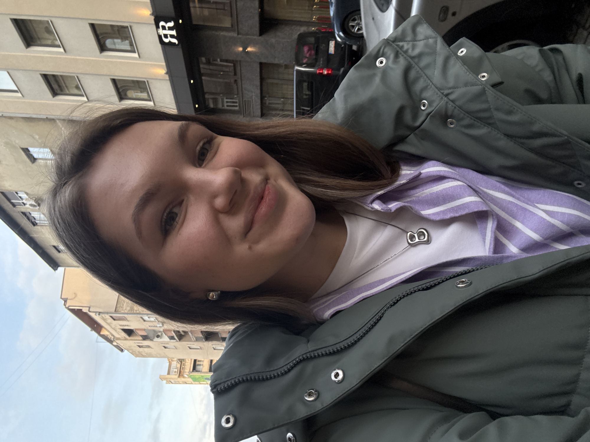 A young woman poses for a selfie outdoors in Belgrade, Serbia. She is wearing a green puffer jacket over a white t-shirt and a purple and white striped shirt. A silver necklace is visible on her chest. Buildings are visible in the background. A black sign with a white symbol on it can be seen on a building behind the woman. A dark-colored vehicle is parked on the street.