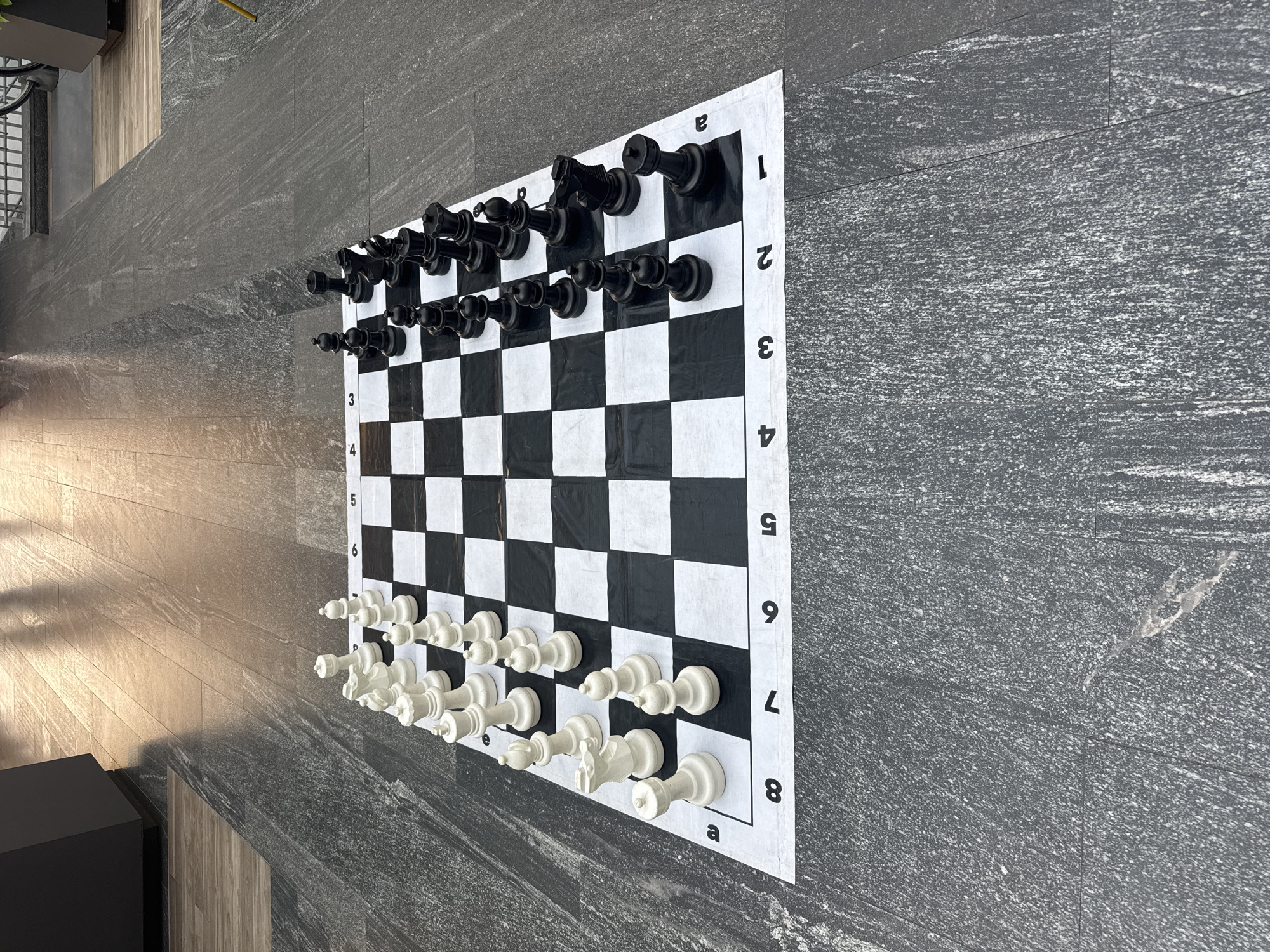 A chessboard with chess pieces set up in the starting position rests on a gray, stone-like surface. The board is large and appears to be made of a black and white patterned material with printed coordinates along the edges. The chess pieces are oversized, one set is black and the other is white. The room is indoors, as evidenced by the presence of a black cabinet or piece of furniture partially visible in the lower left corner and the presence of indoor lighting. The background includes a wall with a stone-like finish, possibly made of large tiles. The scene is static, with no people visible. There is no information to connect it to the city of Puebla, Mexico.