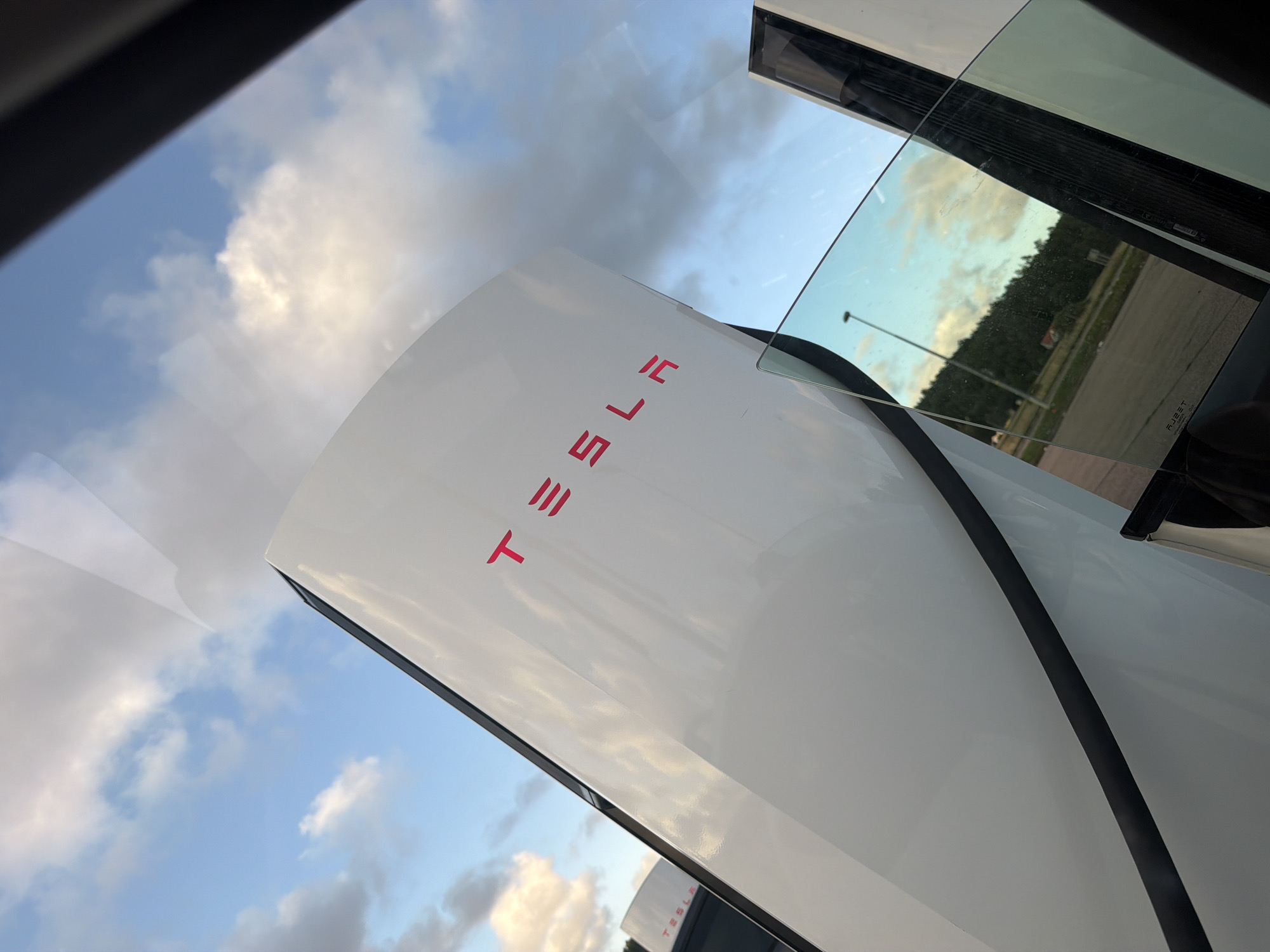 The image captures a close-up view of a Tesla Supercharger station, likely located in Hogstorp, Sweden, given the context. Two white Supercharger units are visible, with the prominent Tesla logo in bright pink lettering on the front of the closer unit. The sky above is filled with a mix of blue and white clouds, illuminated by what appears to be late afternoon or early evening sunlight.
In the foreground, a car window is present, reflecting the sky and a portion of the surrounding environment. Through the tinted glass, a landscape can be seen, featuring a paved road, trees forming a hillside in the distance, and a single lamppost. A small building is also discernible among the trees. The overall scene suggests an outdoor charging location, possibly near a rural or semi-rural area. The angle of the photograph, taken from within a vehicle, implies the photographer is a passenger or driver at the charging station. There are no people visible in the frame.