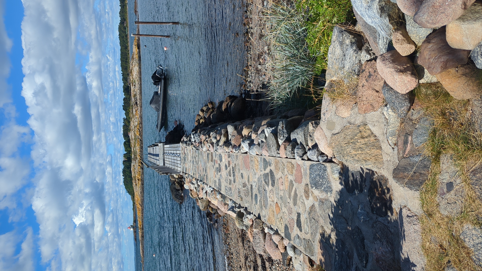 The image depicts a stone pathway leading to a wooden pier extending into the water. The pathway is constructed from various-sized, rough-hewn stones, interspersed with sand. On the right side of the pathway, rocks and dry grass are visible. The wooden pier is visible in the distance, with a black inflatable boat tied to it. The water is calm, with a few buoys floating near the pier. In the background, a rocky island with trees and a lighthouse can be seen under a partly cloudy blue sky. The scene is consistent with a coastal location in Larkollen, Norway, known for its rocky shores and access to the Oslofjord. No people are visible in the image, and no actions or interactions are taking place.