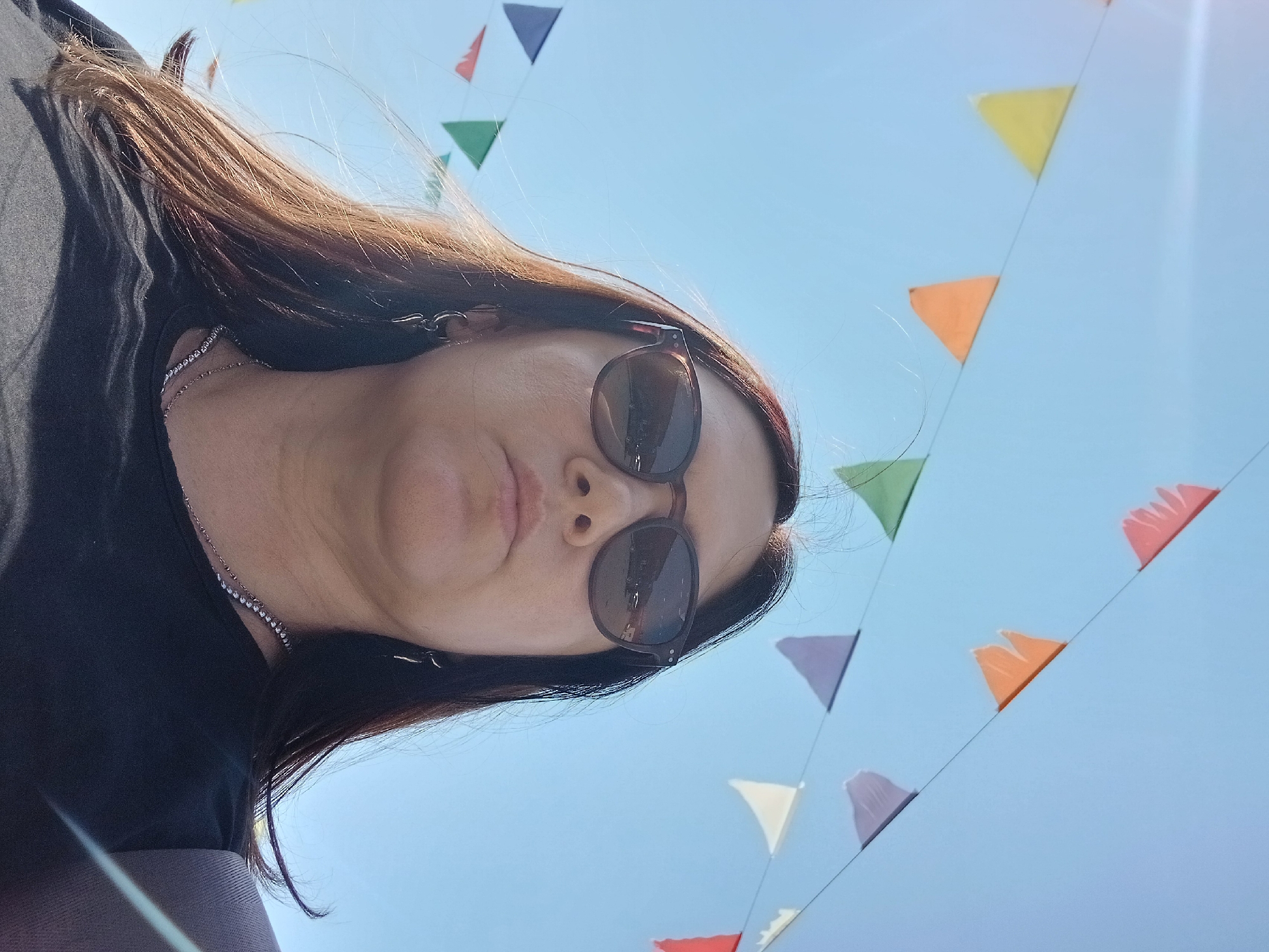 A low-angle, eye-level, medium outdoor shot shows a woman wearing sunglasses and a black t-shirt. She has long, dark brown hair and is wearing a necklace and earrings. Behind her, colorful triangular flags are strung across a clear blue sky. The image is taken in the city of Osa, Russia.