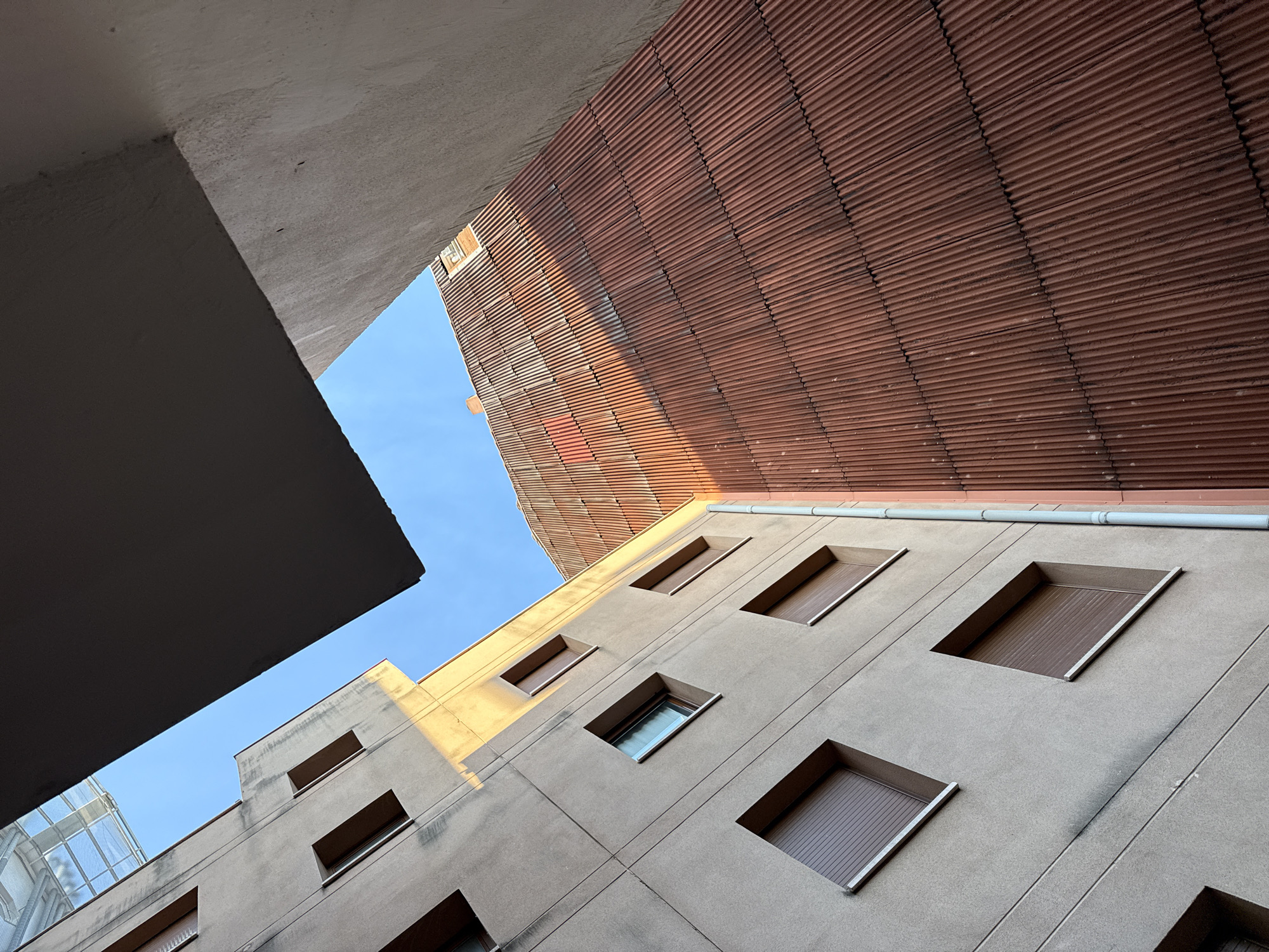 An upward-angled view of buildings in Barcelona, Spain. The composition includes gray concrete buildings with square, recessed windows, some with blinds drawn and some with glass visible. A rusted, corrugated facade is visible above. A corner of a concrete structure in the upper left frames a small section of blue sky. Sunlight casts shadows across the buildings' surfaces. No people are visible.