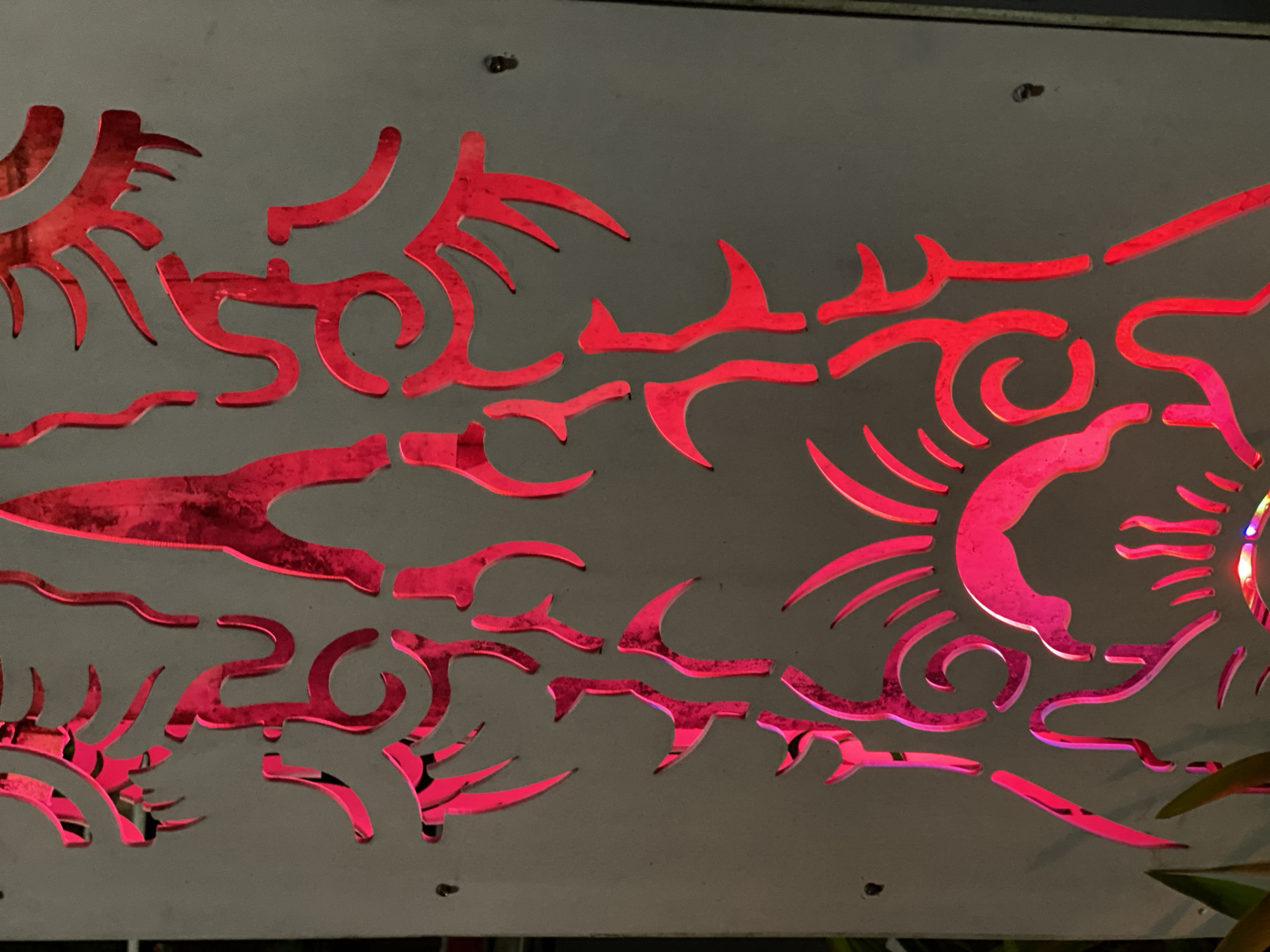 An abstract design is cut into a light gray metal panel, illuminated from behind with bright red light. The design is asymmetrical and appears to be a stylized depiction of organic forms. On the left side, there are shapes resembling flames or stylized claws, as well as curved lines and sharper angles. The center of the panel features a motif that resembles a sun or a burst of energy, with radiating lines and a circular element. The overall impression is one of dynamic movement and vibrant color. The panel is mounted vertically and appears to be part of an outdoor installation, possibly a fence or a decorative screen. There are a few small holes visible in the panel, likely for mounting or ventilation. The background is dark and out of focus, with hints of green foliage on the far left.