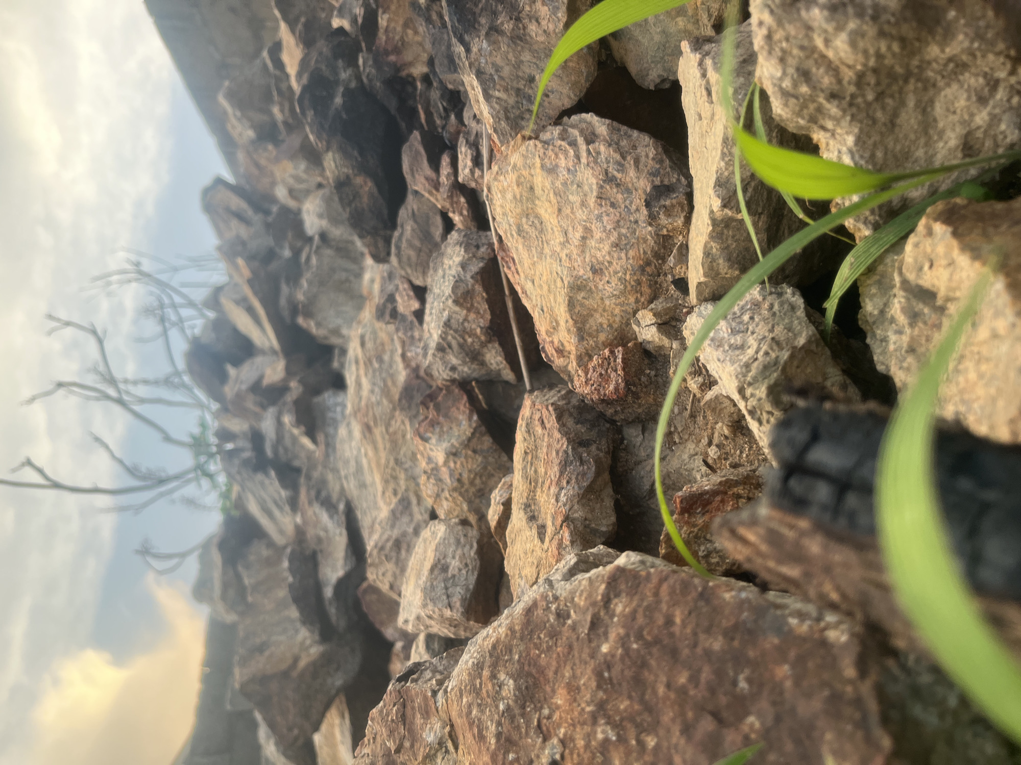 A low-angle shot shows a retaining wall constructed from various sizes of rough-cut rocks. Grass blades and a charred wooden fragment are partially visible among the rocks. The sky is seen in the background. This image was taken in Barakin Akawo, Nigeria.