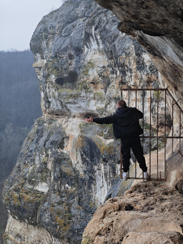 An adult male stands on a narrow, rocky ledge secured by a dark, weathered metal barrier with vertical bars, situated within a large rock formation near Shumen, Bulgaria. He is viewed from the rear, wearing a dark jacket, dark trousers, and white athletic shoes. His left arm is extended horizontally outwards, palm open, towards a towering, multi-colored rock cliff that dominates the left and central portion of the frame. The background reveals a distant, hazy forested valley under an overcast sky. The immediate foreground includes the rough texture of a rocky overhang above and to the right of the subject's position.