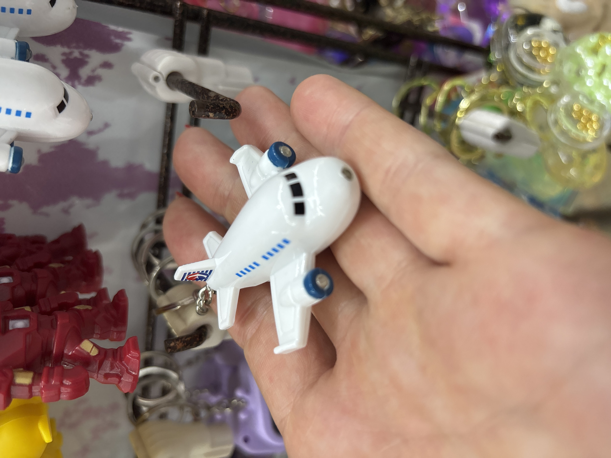 A close-up, eye-level shot shows a person's palm holding a small, white toy airplane keychain with blue accents. The airplane has two engines and a tail with a logo. In the background, out of focus, are various other small toys and keychains, including red robot figures, a yellow object, and colorful, metallic-looking trinkets. The overall impression is of a display of small collectible items. The image does not contain any information about the location of Sochi, Russia.