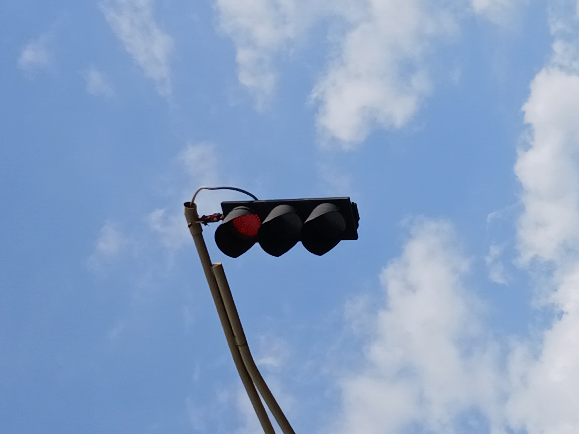 An image captured in Ciudad Guayana, Venezuela, showcases a traffic light set against a bright blue sky with scattered white clouds. The primary subject is a horizontal traffic light, which has its leftmost signal illuminated red, indicating a stop. The other two signal lights are dark.
The traffic light is mounted on a light-colored, possibly beige or brown, metal pole that extends diagonally upwards from the lower left of the frame. Some electrical wiring is visible near the connection point of the traffic light to the pole. The perspective is from below, looking up at the traffic signal.
The background is a clear, vibrant blue sky, suggesting fair weather. Several fluffy white clouds are distributed across the sky, adding texture to the otherwise smooth blue expanse. The lighting indicates it is daytime, likely a bright and sunny day. There are no people, vehicles, or buildings visible in the frame, focusing solely on the traffic light and the sky. No discernible text is present in the image.