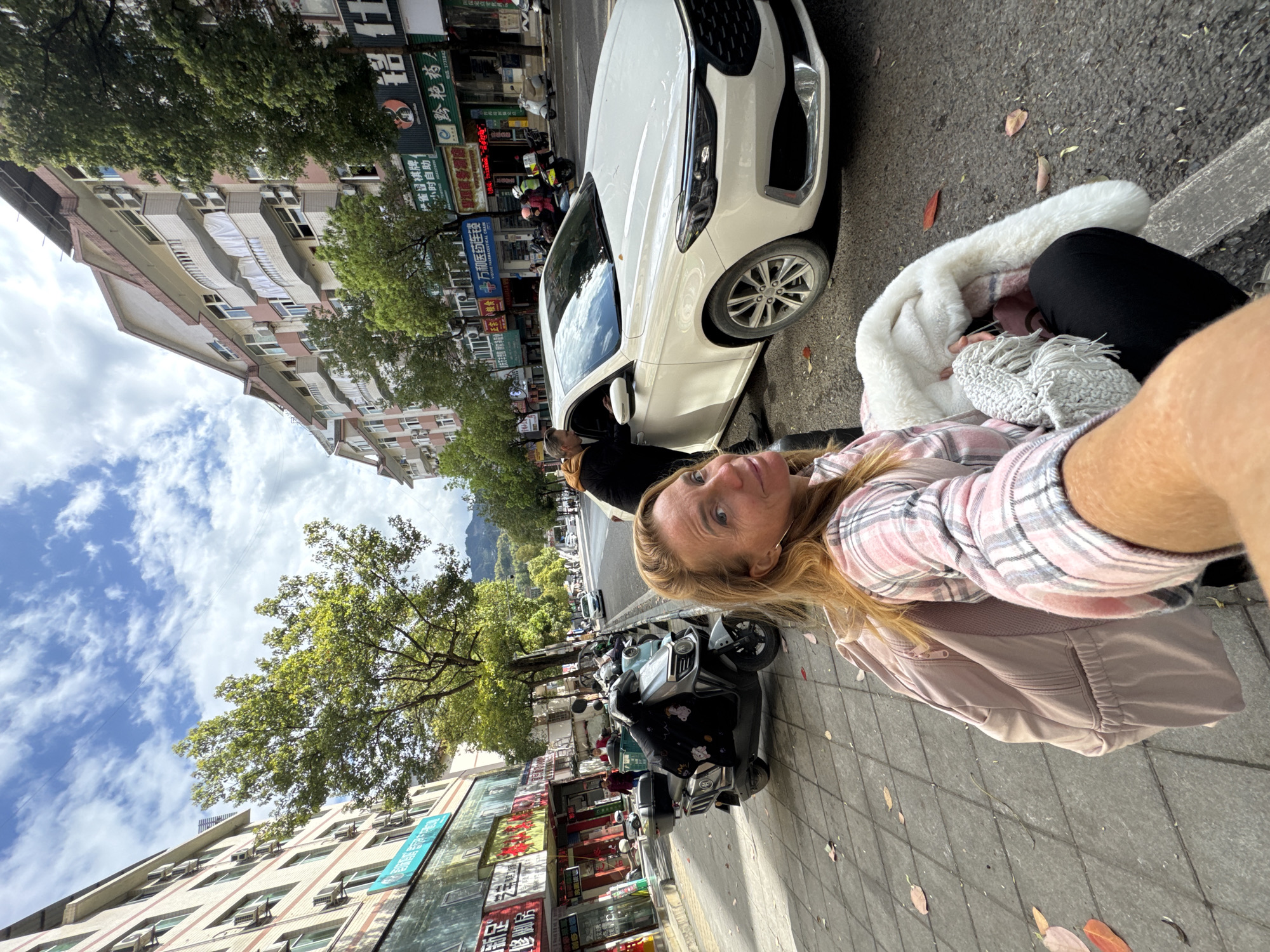 The photograph captures a woman taking a selfie on a street in Qianjiang, China. The woman, positioned on the right side of the image, is wearing a pink and white plaid shirt and a pink jacket. To her left is a white car parked on the side of the road, with another person visible in the driver's seat. The background features a typical urban street scene, with buildings displaying Chinese characters, trees lining the street, and various parked scooters and vehicles. The sky above is partly cloudy.