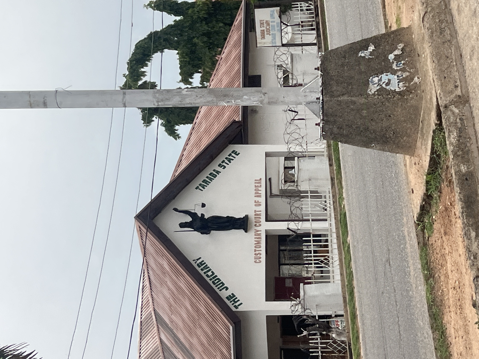 The image shows the Customary Court of Appeal building in Jalingo, Taraba State, Nigeria. The building is white with a brown, corrugated metal roof. Above the entrance, a statue of Lady Justice stands in front of text that reads "THE JUDICIARY TARABA STATE". Below the statue, "CUSTOMARY COURT OF APPEAL" is written in red letters. A white fence with barbed wire on top encloses the property. A grey utility pole stands in the foreground, with its base resting on a concrete structure adorned with torn posters. A street runs in front of the building, and the ground on either side of the street is covered with dirt and sparse vegetation. The sky is overcast.