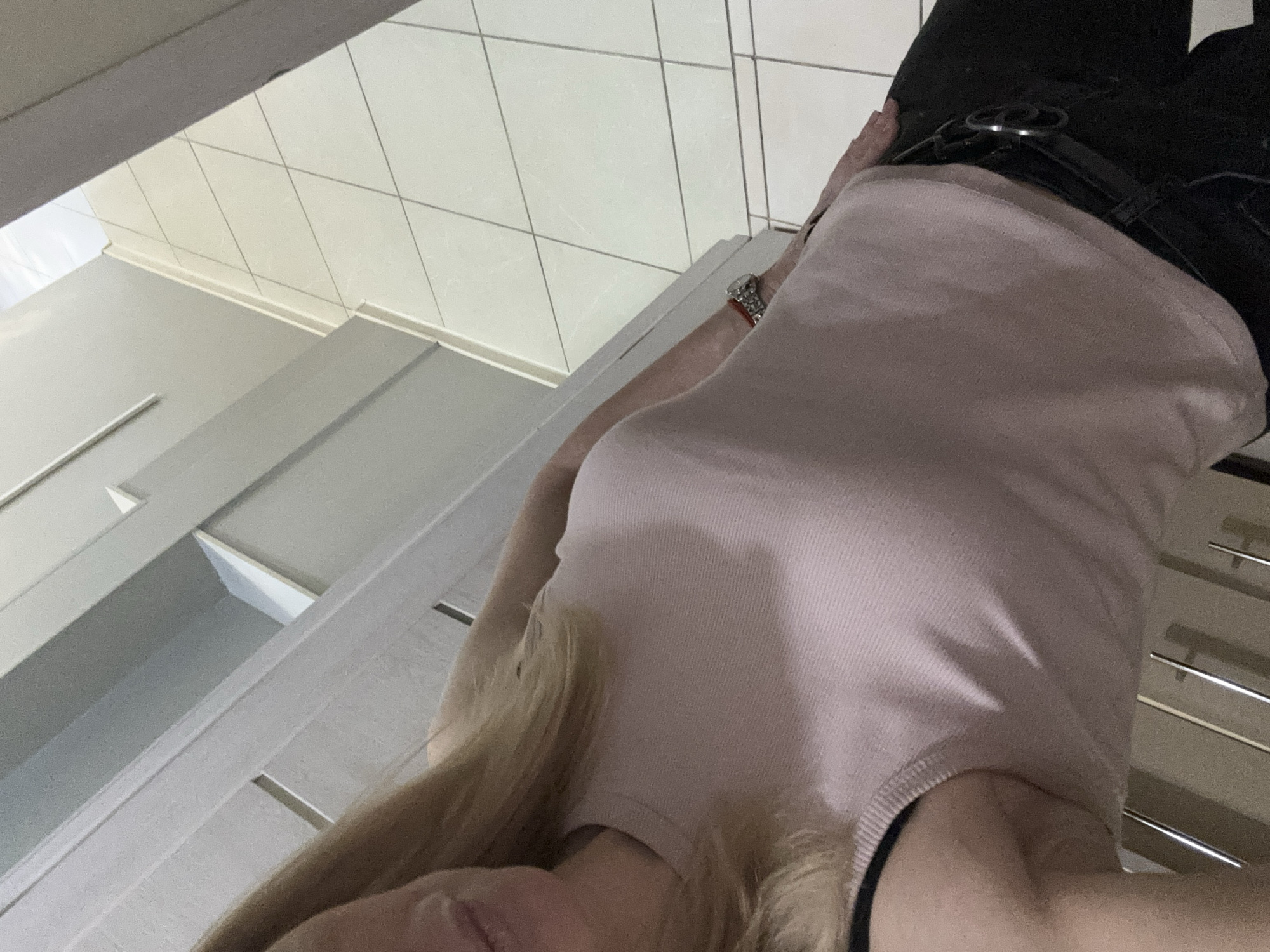 The image shows a woman wearing a light pink tank top and black pants with a belt, likely in an indoor setting. A watch is visible on her wrist. The background includes a white tile floor and a light-colored cabinet or shelving unit. The location appears to be inside a building in the city of Vsevolozhsk, Russia.
