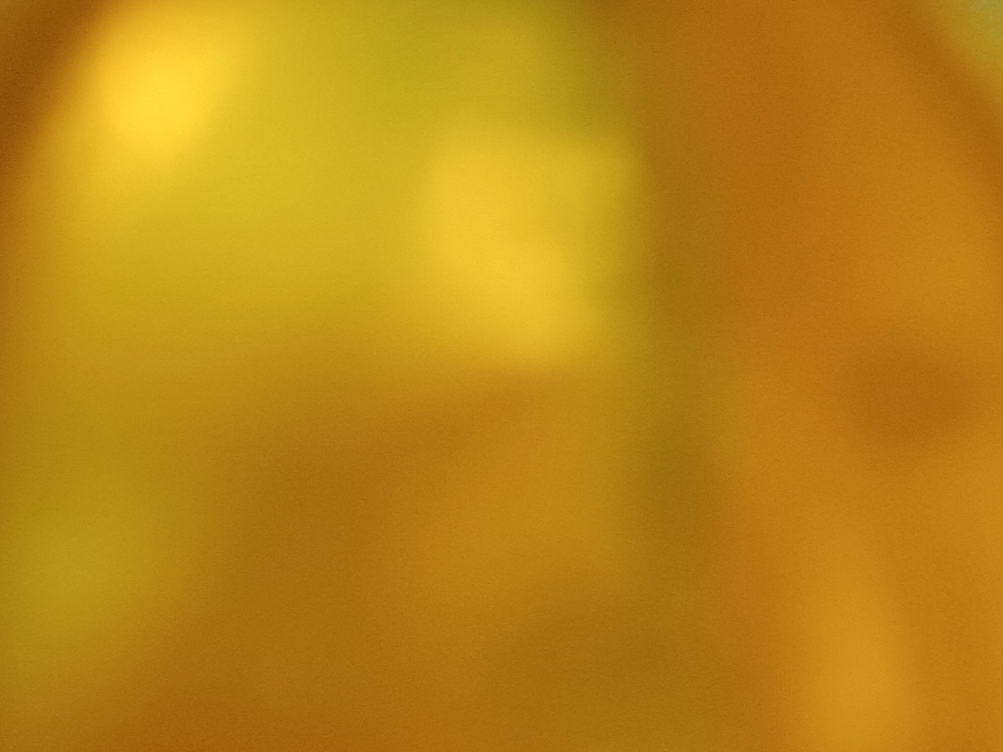 This is an abstract, out-of-focus image with a dominant golden-yellow and orange gradient. There are no discernible subjects, objects, or a specific scene depicted. The lighting creates soft, blurry areas of brightness and shadow across the image, with a brighter, more yellow area in the upper left quadrant fading into a warmer, more orange tone towards the right and bottom. The texture appears grainy, suggesting it may be a photograph. There are no visible text elements, location cues, or indications of time of day or weather. The image lacks any discernible activity or event, and therefore, no emotions can be inferred. Given the location context of Zaporizhzhia, Ukraine, and the abstract nature of the image, it is not possible to provide any specific contextual details beyond the visual characteristics of the image itself.
