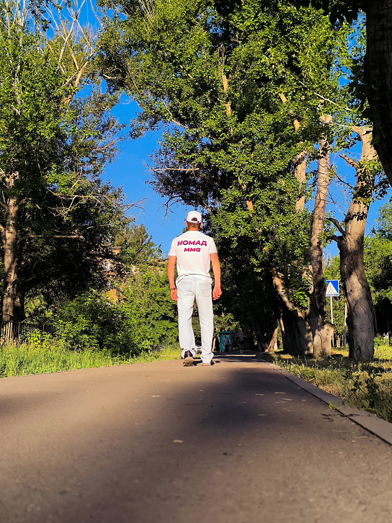 A man wearing a white t-shirt with Russian text and white pants walks away from the camera on a paved path lined with trees and green foliage under a clear blue sky. The t-shirt has red text that appears to read "НОМАД ММО". The path is surrounded by lush greenery and large trees. In the background, a building is partially visible through the trees, and a pedestrian crossing sign can be seen on the right side of the path. The sun casts a shadow from the trees onto the path. The scene suggests a warm, sunny day in a park-like setting within the city of Qarağandı, Kazakhstan.