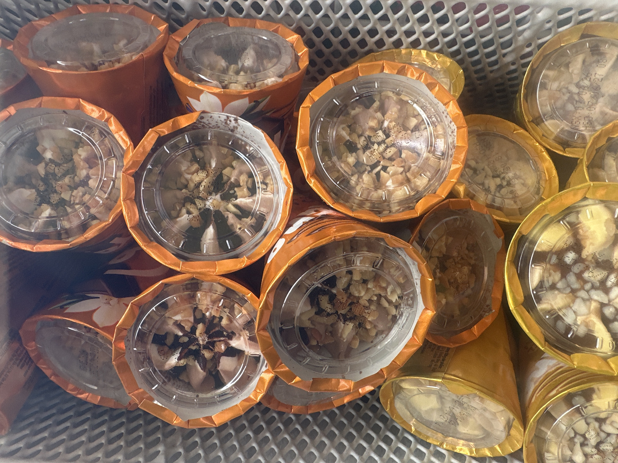 This is a close-up, top-down view of a collection of individual ice cream cups, likely arranged in a container or display. The cups are primarily orange and yellow, with clear plastic lids. Many of the cups are filled with a light-colored substance, possibly ice cream, topped with chopped nuts and what appears to be a dusting of cinnamon or chocolate powder. Some cups show visible text and dates printed on their lids or wrappers, suggesting a retail or commercial setting. The overall impression is of a neatly organized display of single-serving desserts. The location is described as Bac Nha Trang Ward, Vietnam, implying this scene is from a food establishment or market in that area. There are no people visible in the image.