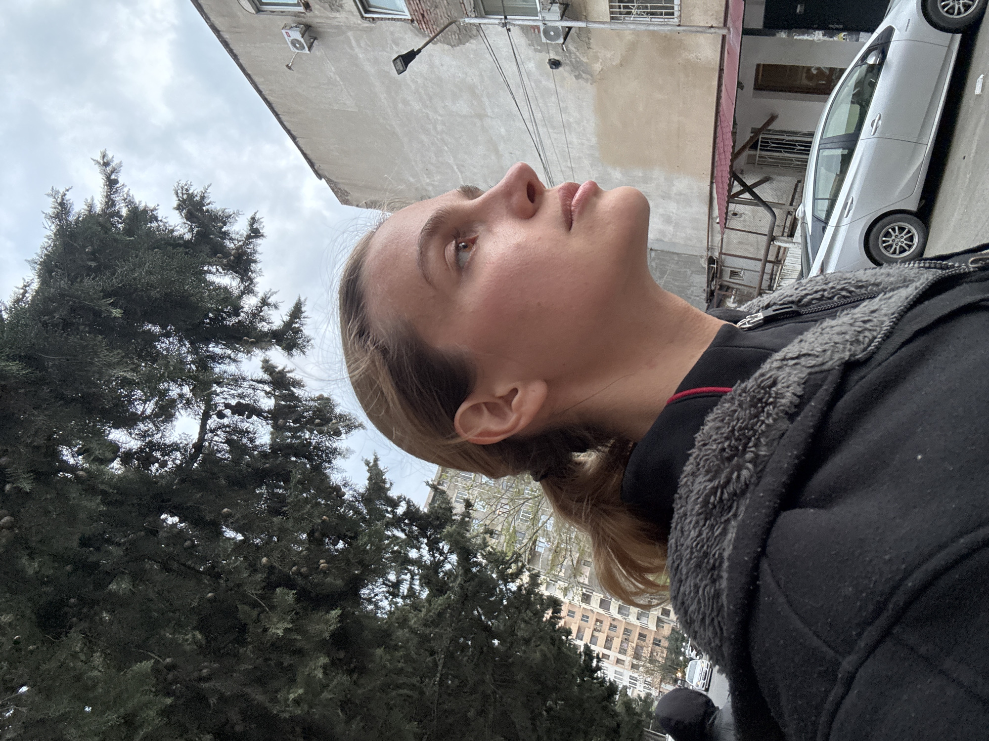 The image presents an upward-looking view in an urban setting, contextually identified as Tbilisi, Georgia. In the immediate foreground, the right profile of a young adult's face and upper body is visible. She has light-colored hair, appearing to be pulled back, and wears a black jacket with a distinct grey, fluffy collar. Her gaze is directed upwards and slightly to the right, with a neutral, contemplative expression.
The sky above is overcast, characterized by light grey clouds, indicating a cloudy day with soft, diffuse natural light. To the left, several dark green coniferous trees, possibly cypress or juniper, are prominent, their branches densely packed with numerous small, round cones.
Further into the background, older, textured buildings with concrete or stucco facades are visible. These structures feature multiple windows, some fitted with grilles. One building has a street lamp extending from its side, and another displays a small, white outdoor air conditioning unit. On the right side of the frame, a silver or light-colored sedan, possibly a Toyota Prius, is parked along what appears to be a street. The entire image is captured with a tilted perspective, offering a dynamic snapshot of an ordinary urban moment. No discernible text is visible.