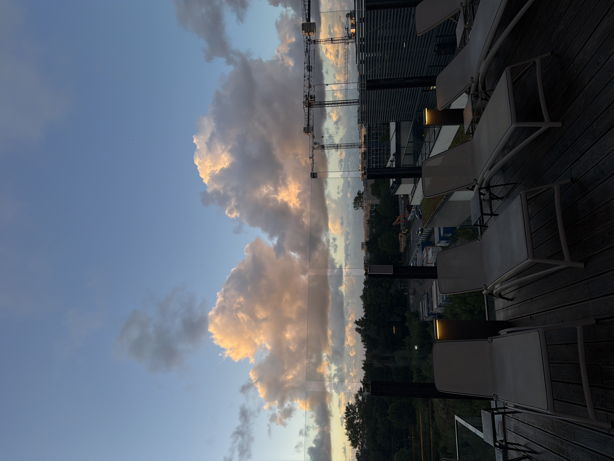 The image depicts a rooftop scene at sunset in Kołobrzeg, Poland, overlooking a landscape of trees, construction cranes, and modern buildings. In the foreground, several empty lounge chairs are arranged on a wooden deck. A glass railing separates the deck from the view beyond. The sky is filled with dramatic, illuminated clouds, suggesting the end of the day. The overall impression is one of urban development against a natural backdrop, bathed in the warm light of sunset.