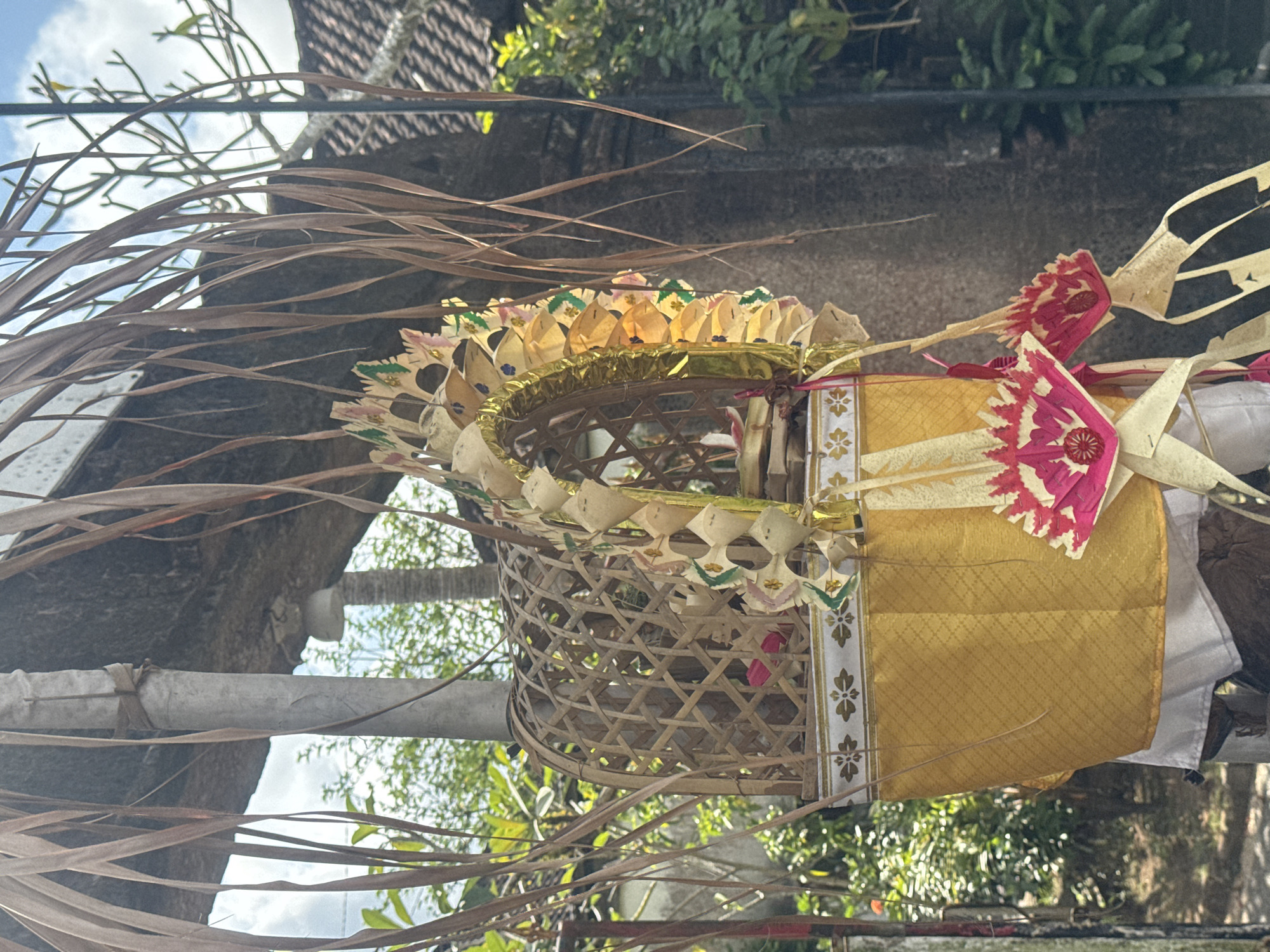 The image depicts an outdoor scene in Kuta Selatan, Indonesia, featuring traditional Balinese offerings or decorations. The foreground is dominated by a woven bamboo structure, possibly a basket or a decorative element, adorned with yellow fabric and intricate paper cuttings of pink and yellow flowers. Dried palm fronds hang from above, framing the top of the image. In the background, lush green foliage and trees are visible, along with glimpses of traditional Indonesian architecture, including tiled roofs and stone structures. The scene suggests a cultural or religious setting, possibly related to a ceremony or temple.