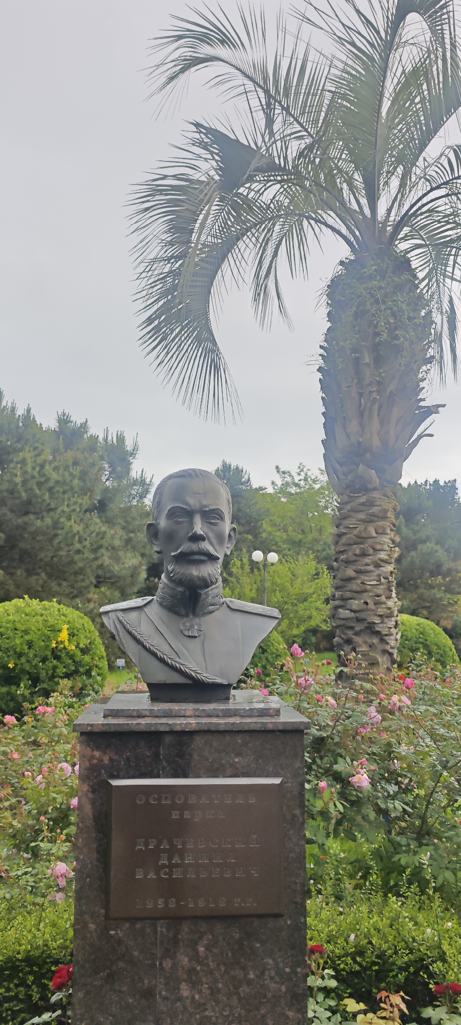 The image depicts a bust of a man in a park setting. The bust is positioned on a dark granite pedestal with a plaque that reads: "Основатель Парка. Драчевский Даниил Васильевич. 1858-1918 г.г." which translates to "Founder of the Park. Drachevsky Daniil Vasilyevich. 1858-1918". The bust shows a man with a mustache and beard, wearing what appears to be a military uniform with epaulets and a sash. The background includes a palm tree, other trees and bushes, and a flowerbed filled with pink roses.
Based on the plaque, the location is in a park founded by Daniil Vasilyevich Drachevsky. Given the Cyrillic text on the plaque, it's plausible that the location could be in Russia. Further research would be needed to confirm if this park is in the city of Sirius, Russia.